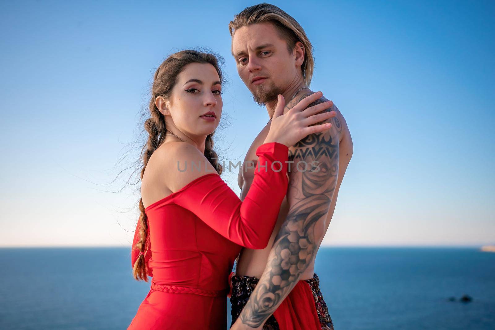 Portrait of tattooed man and sensual young brunette woman in red dress, happily dancing outdoors isolated on blurry sea background with natural bokeh in soft warm sunset backlight. Selective focus. by panophotograph