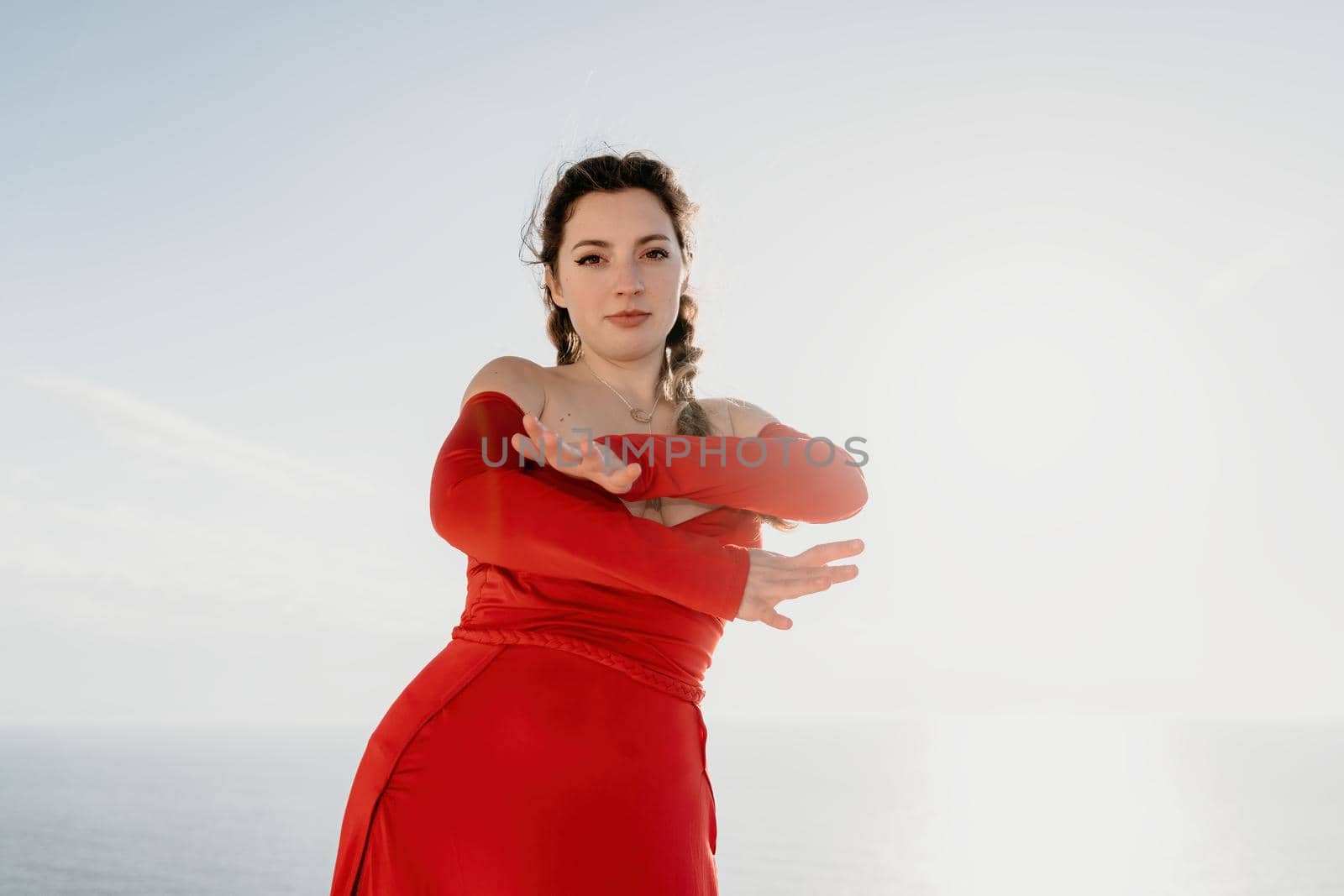 Closeup portrait of sensual young brunette woman in red dress, happily dancing outdoors isolated on blurry sea background with natural bokeh in soft warm sunset backlight. Selective focus. by panophotograph