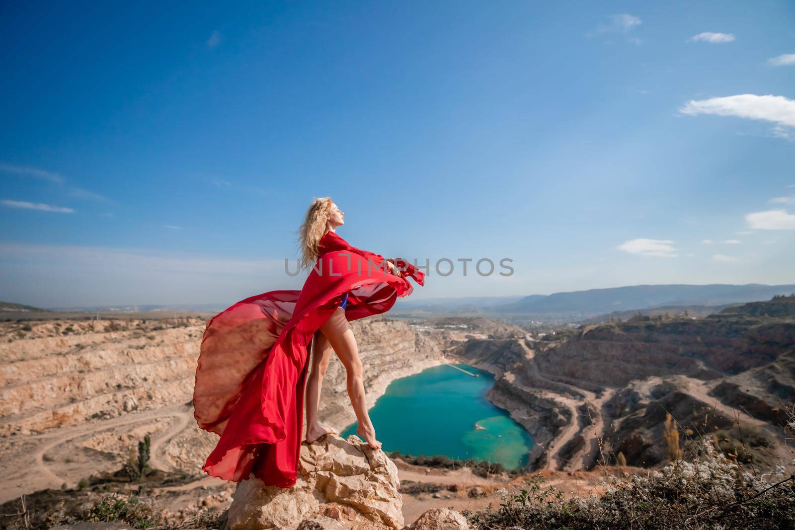 Side view of a beautiful sensual woman in a red long dress posing on a rock high above the lake in the afternoon. Against the background of the blue sky and the lake in the form of a heart by Matiunina