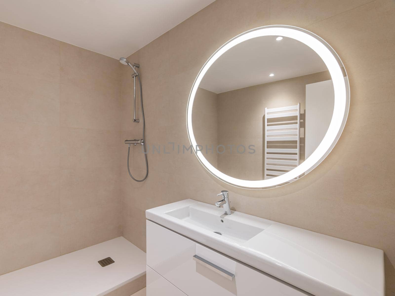 Modern bathroom with beige tiles, furniture, shower and round large mirror with lighting.