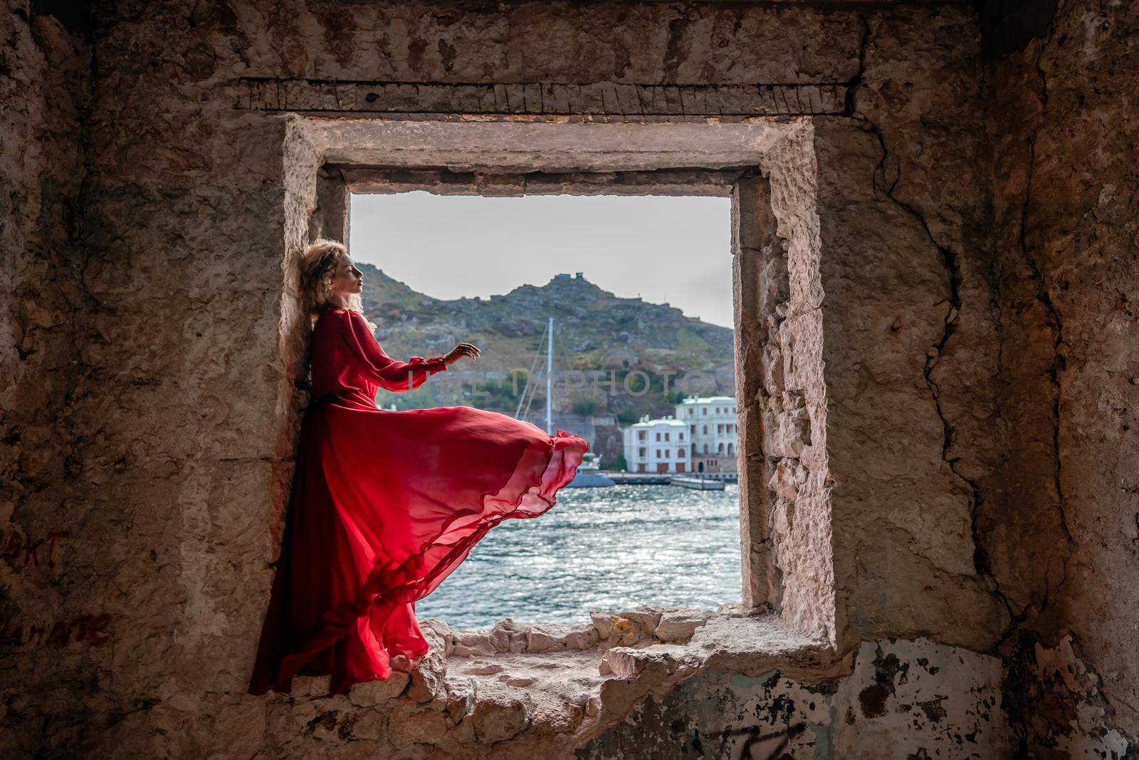 View of Balaklava Bay through an arched balcony in oriental style. The girl in a long red dress stands with her back. Abandoned mansion on the Black Sea coast.