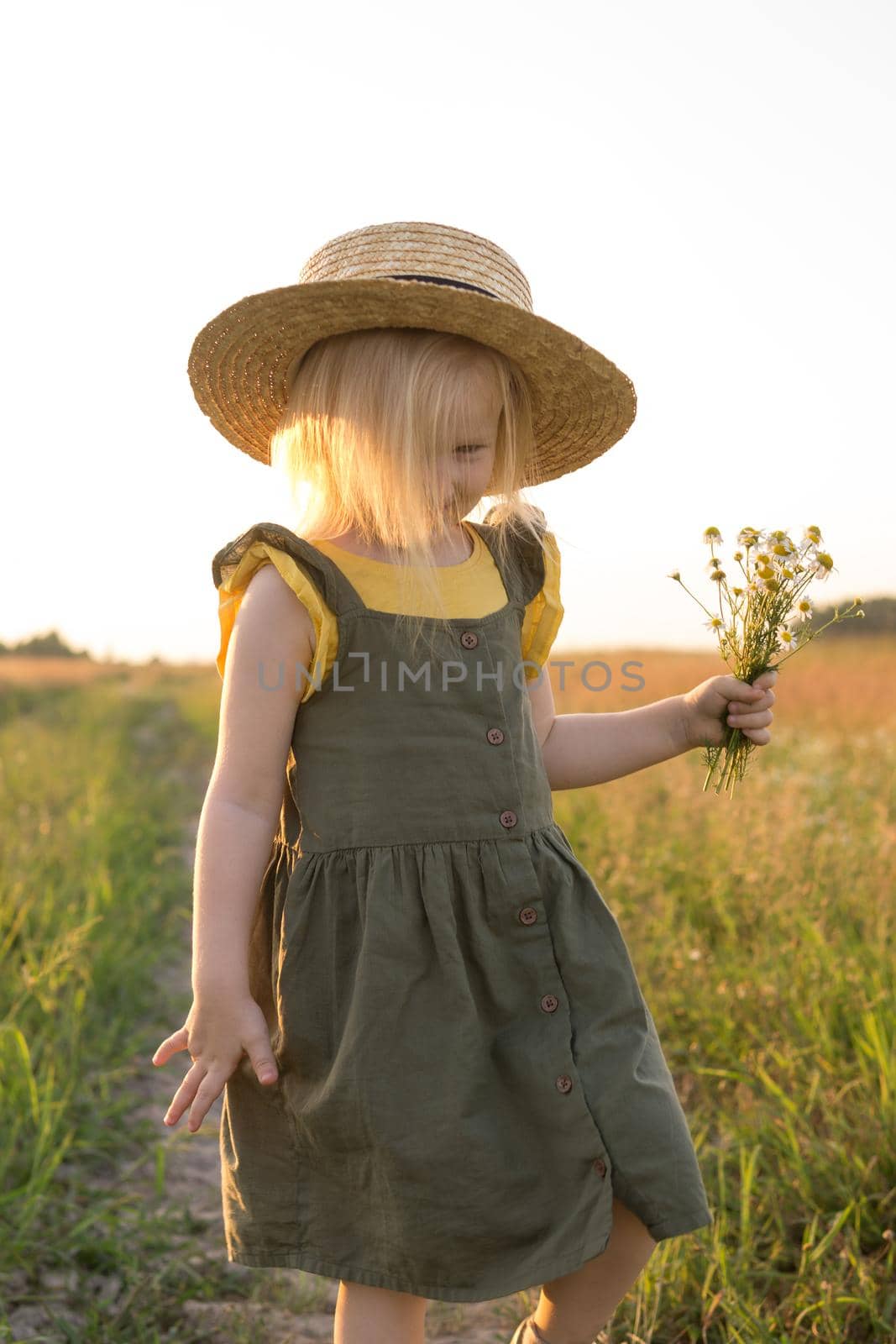 A little blonde girl in a straw hat walks in a field with a bouquet of daisies. The concept of walking in nature, freedom and an eco-friendly lifestyle