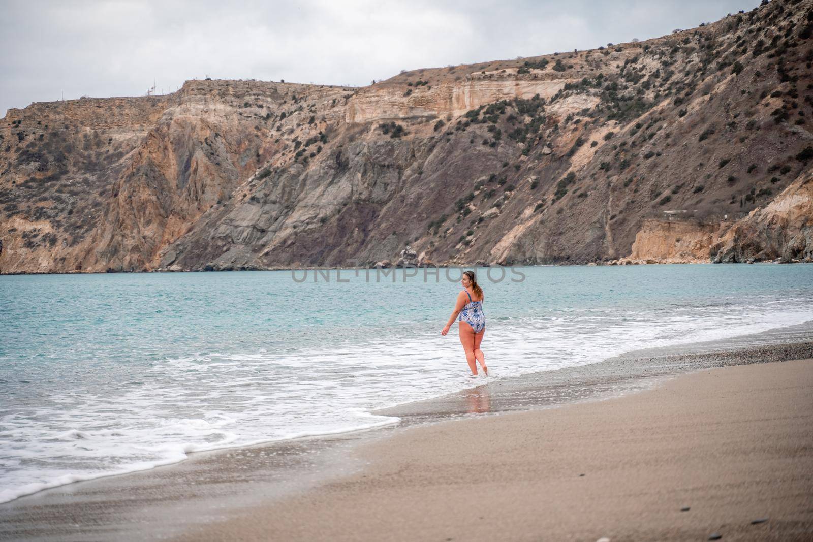 A plump woman in a bathing suit enters the water during the surf. Alone on the beach, Gray sky in the clouds, swimming in winter