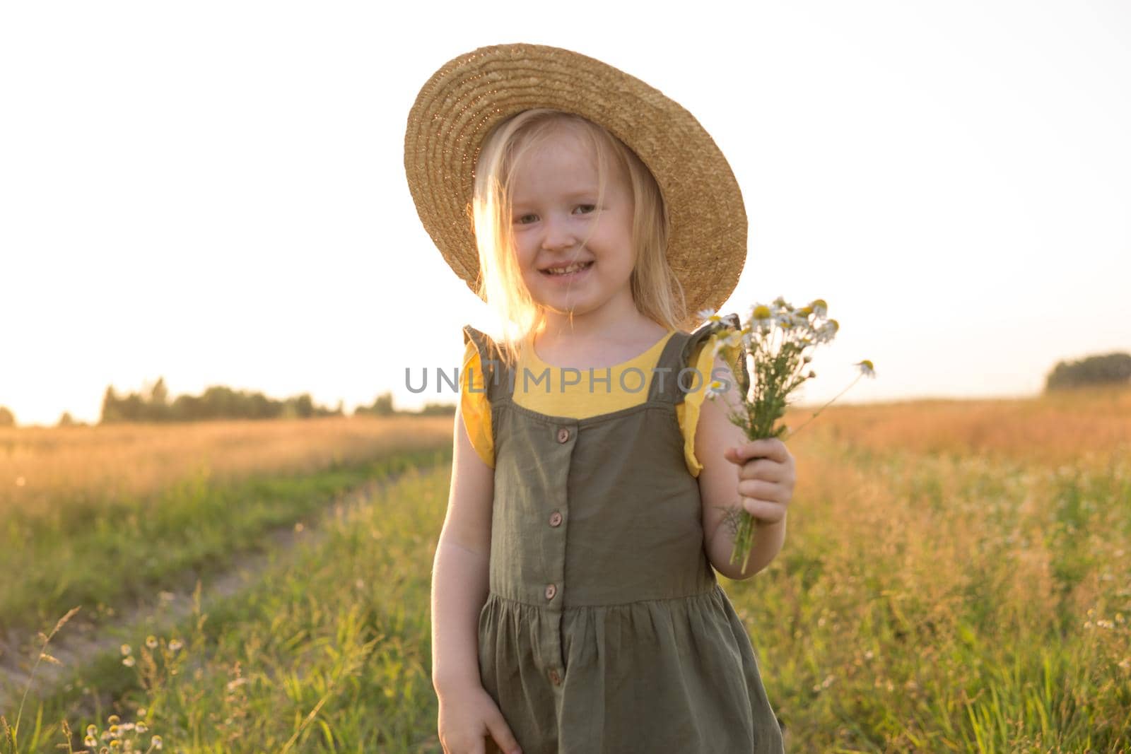 A little blonde girl in a straw hat walks in a field with a bouquet of daisies. The concept of walking in nature, freedom and an eco-friendly lifestyle.