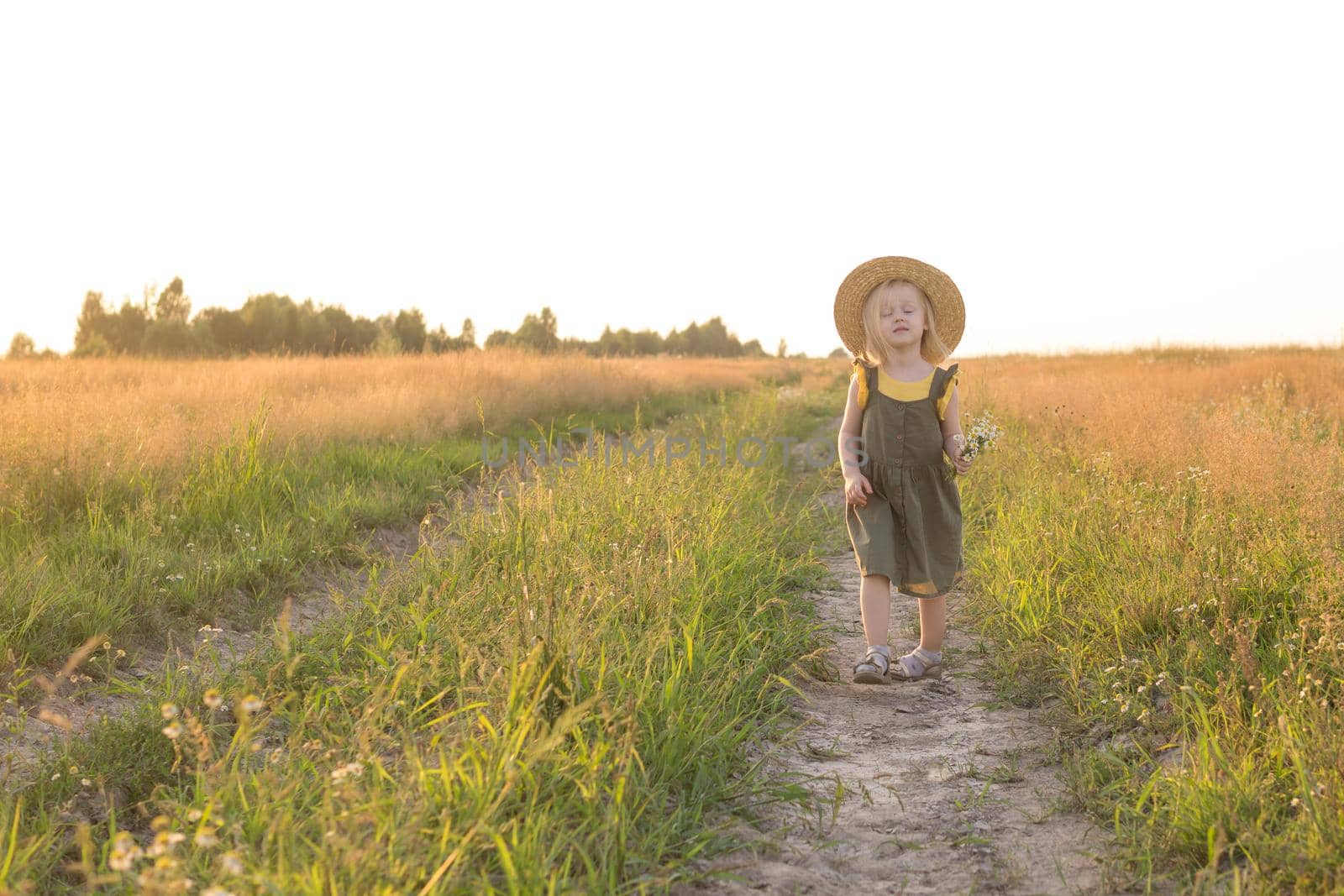 A little blonde girl in a straw hat walks in a field with a bouquet of daisies. The concept of walking in nature, freedom and an eco-friendly lifestyle.