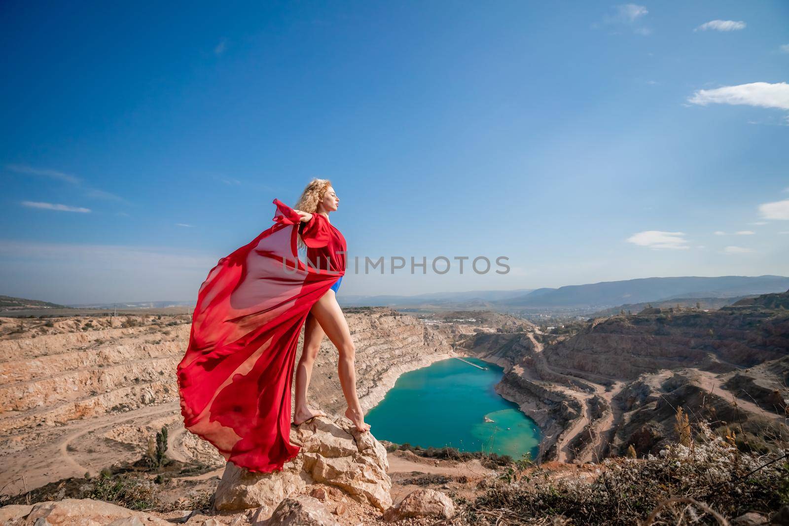 Side view of a beautiful sensual woman in a red long dress posing on a rock high above the lake in the afternoon. Against the background of the blue sky and the lake in the form of a heart.