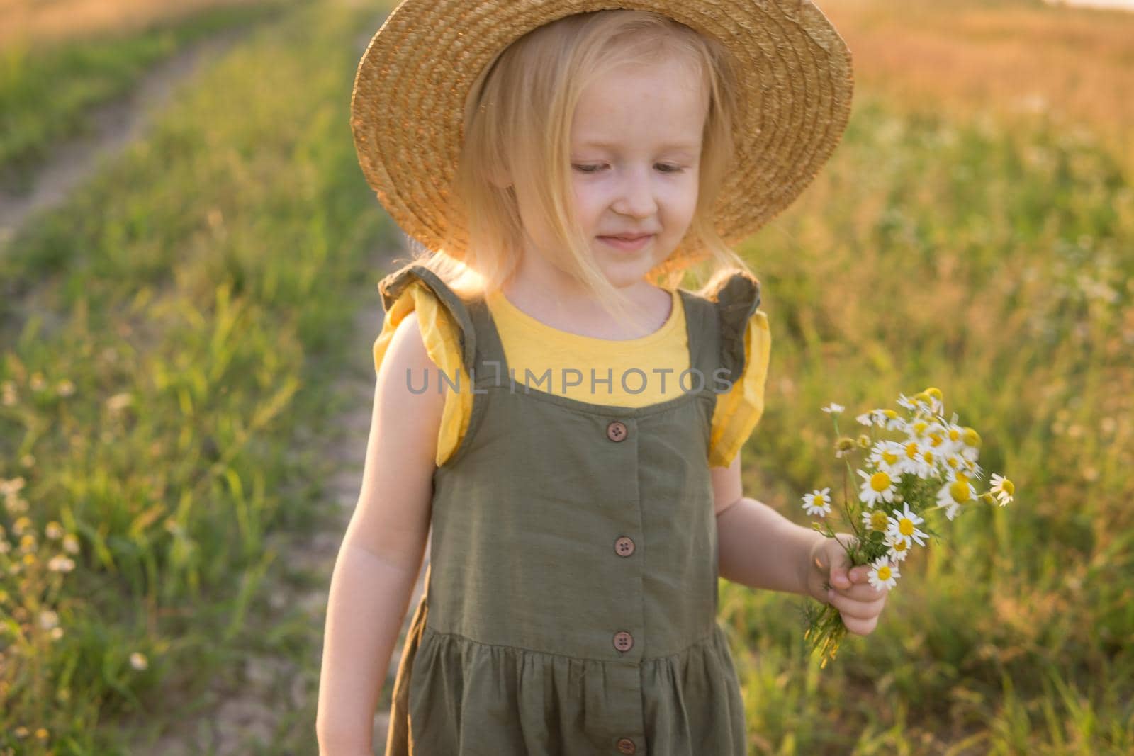 A little blonde girl in a straw hat walks in a field with a bouquet of daisies. The concept of walking in nature, freedom and an eco-friendly lifestyle.