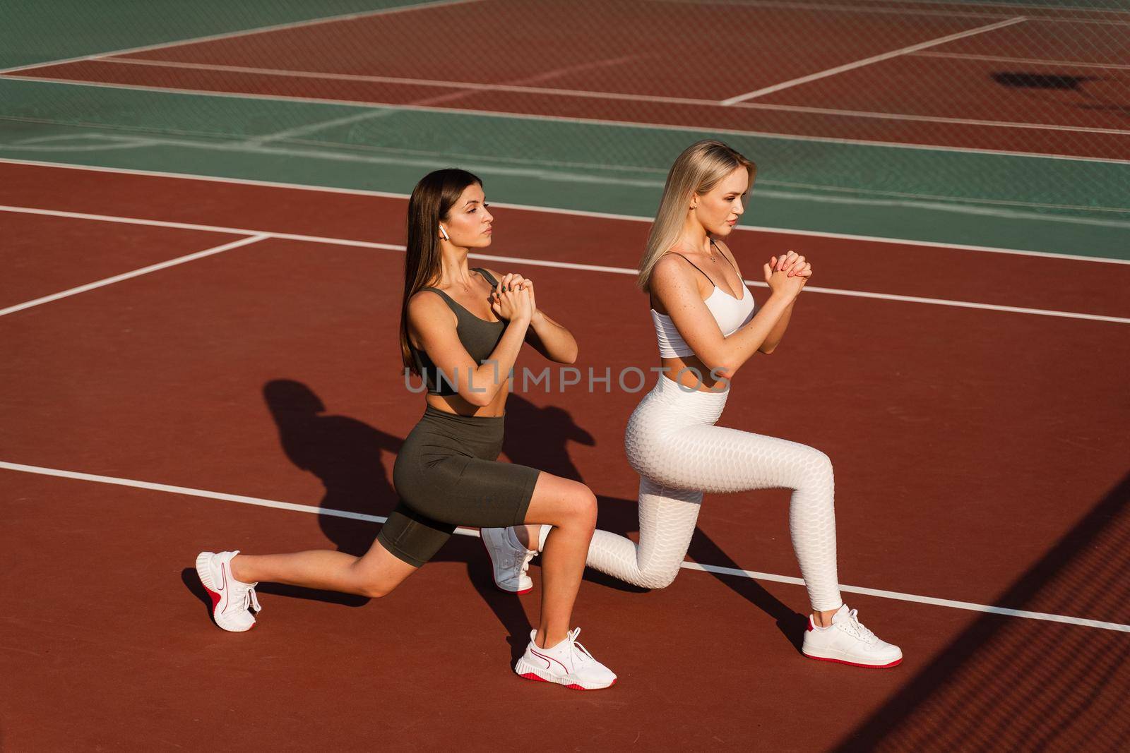 2 girls doing lunges training for stretching legs. Sport exercises on the tennis court