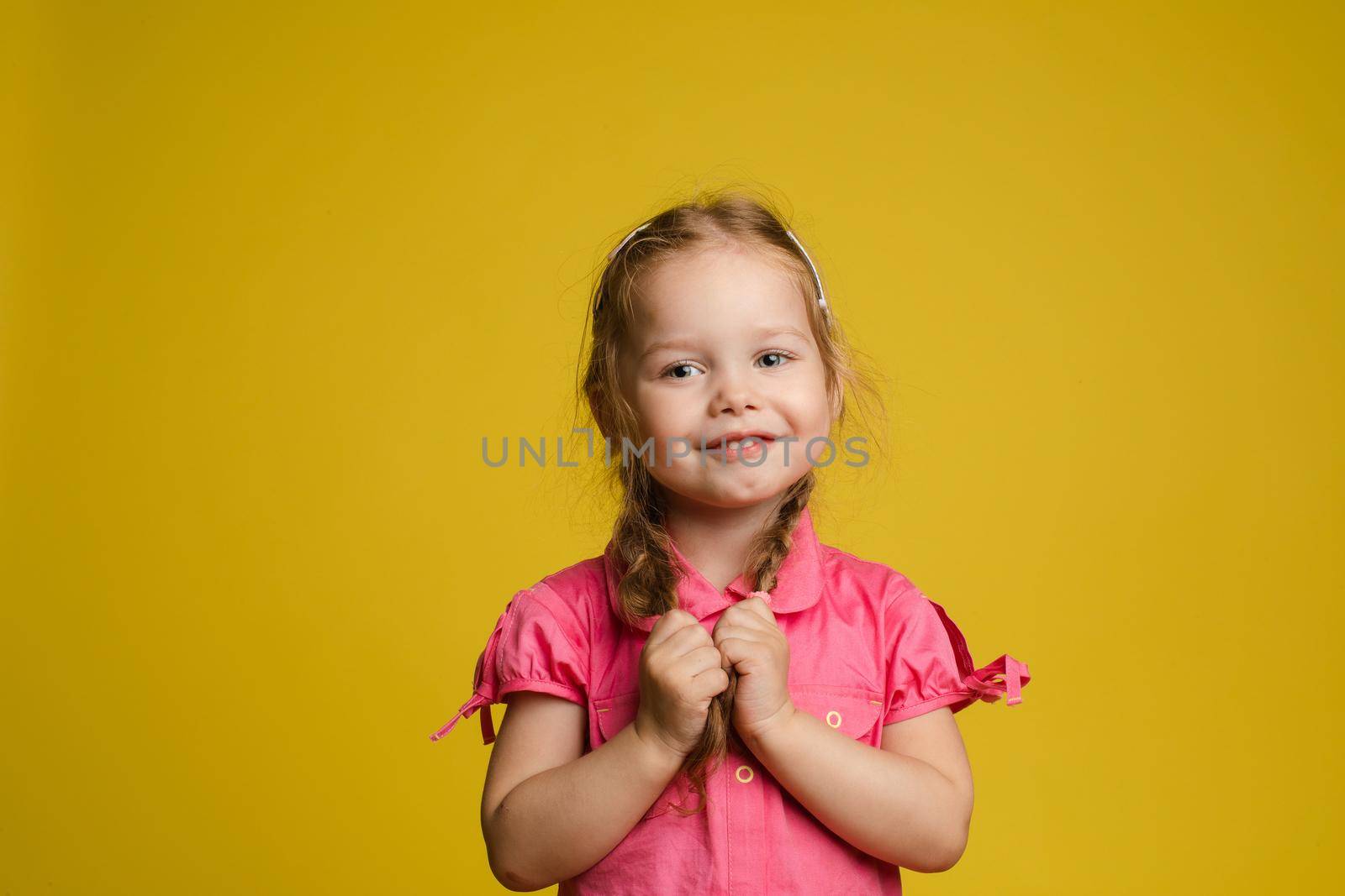 Portrait of adorable little cute female child holding hair posing looking at camera medium shot. Smiling pleasant happy girl having positive emotion isolated at yellow studio background