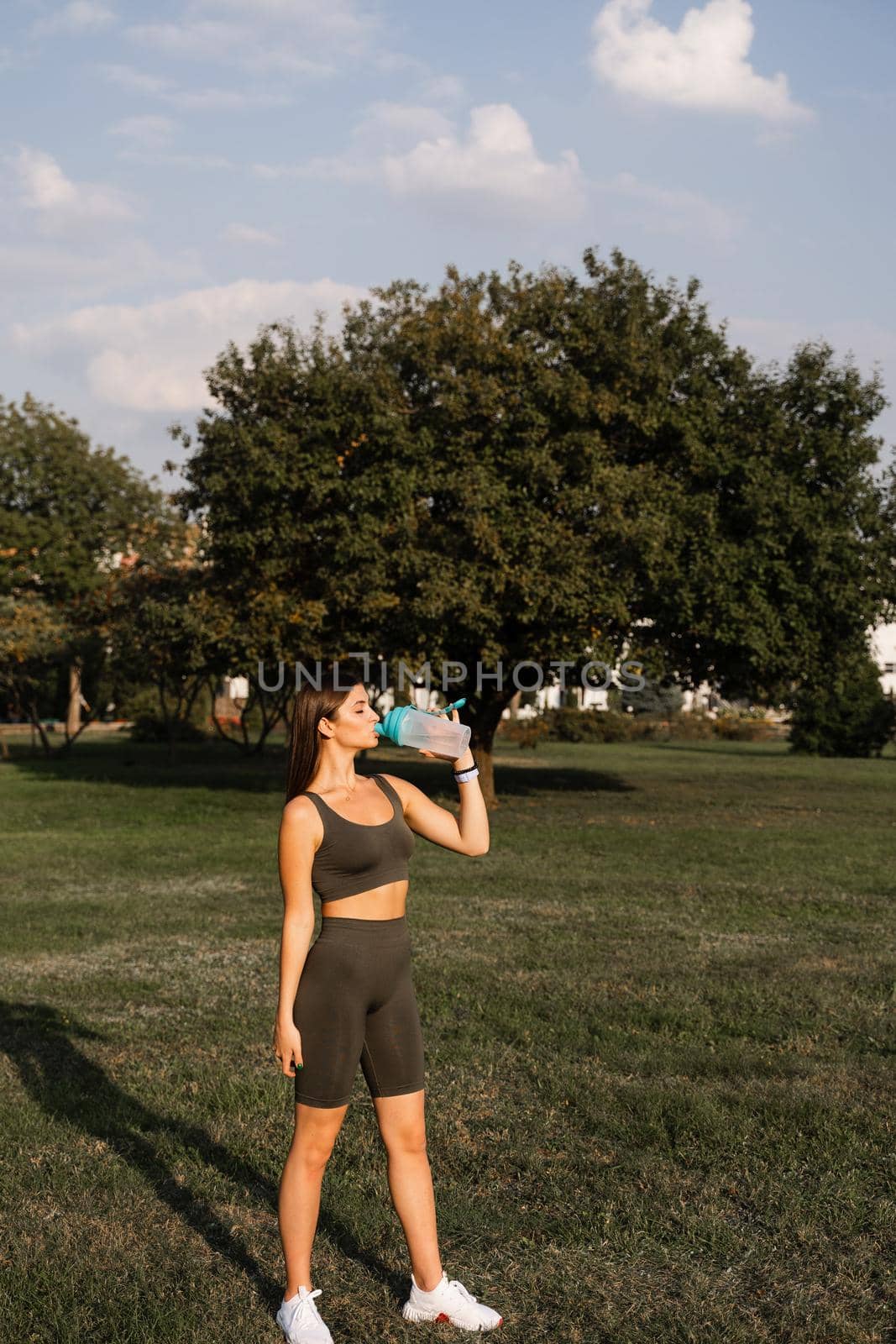 Athletic fit Asian girl drinking water from a bottle in green park. Rest after training. by Rabizo