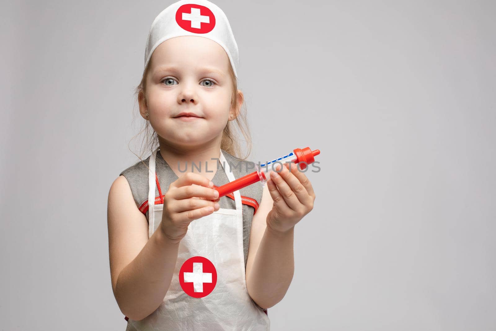 Little cute funny girl playing wearing doctor uniform holding toy syringe looking at camera. Smiling baby female child in glasses posing in nurse garb at white studio background medium long shot