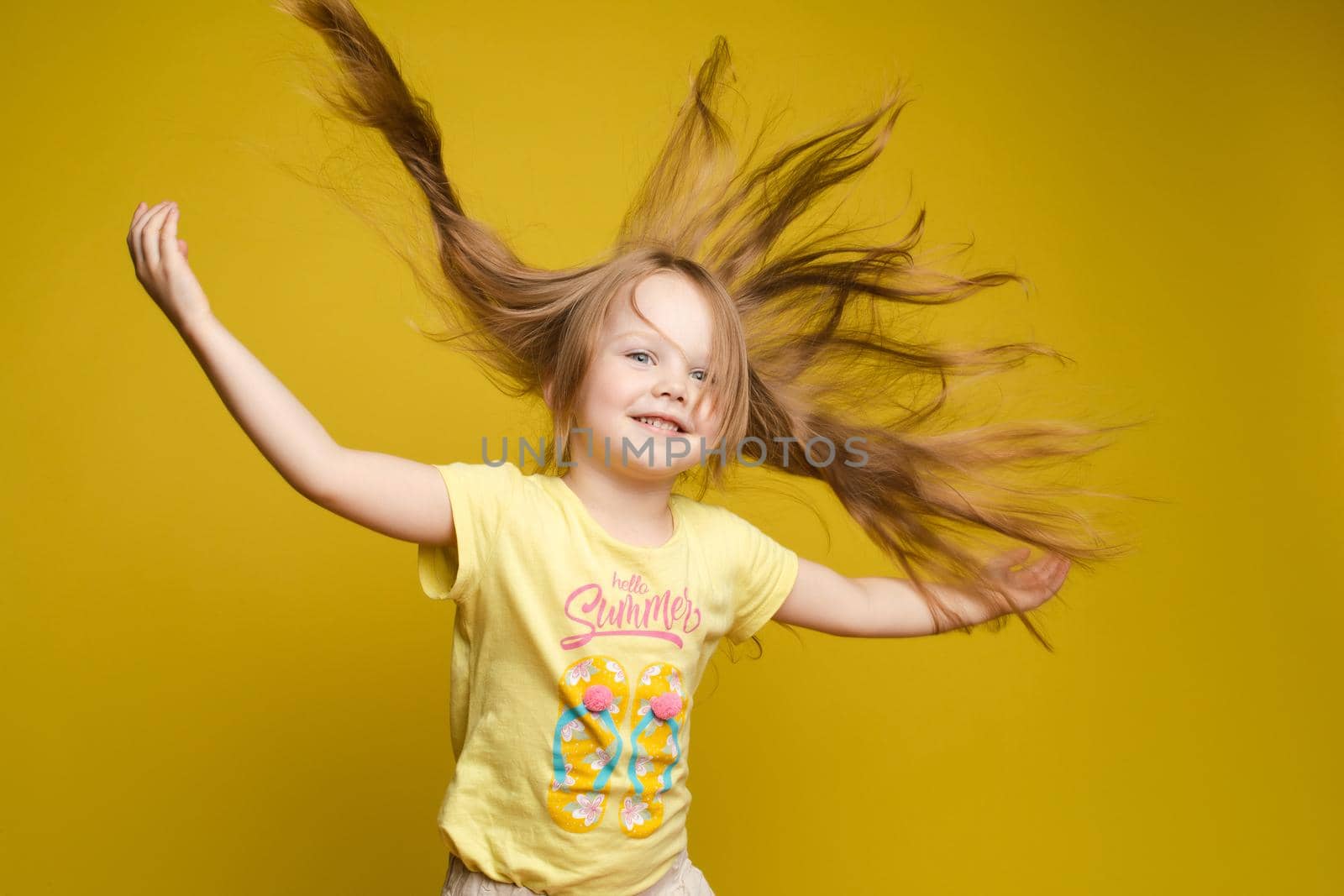 Front view of beautiful longhaired girl in cute shirt playing with hair and twirling in studio. Sunny little child looking at camera and posing on yellow isolated background. Concept of childhood.