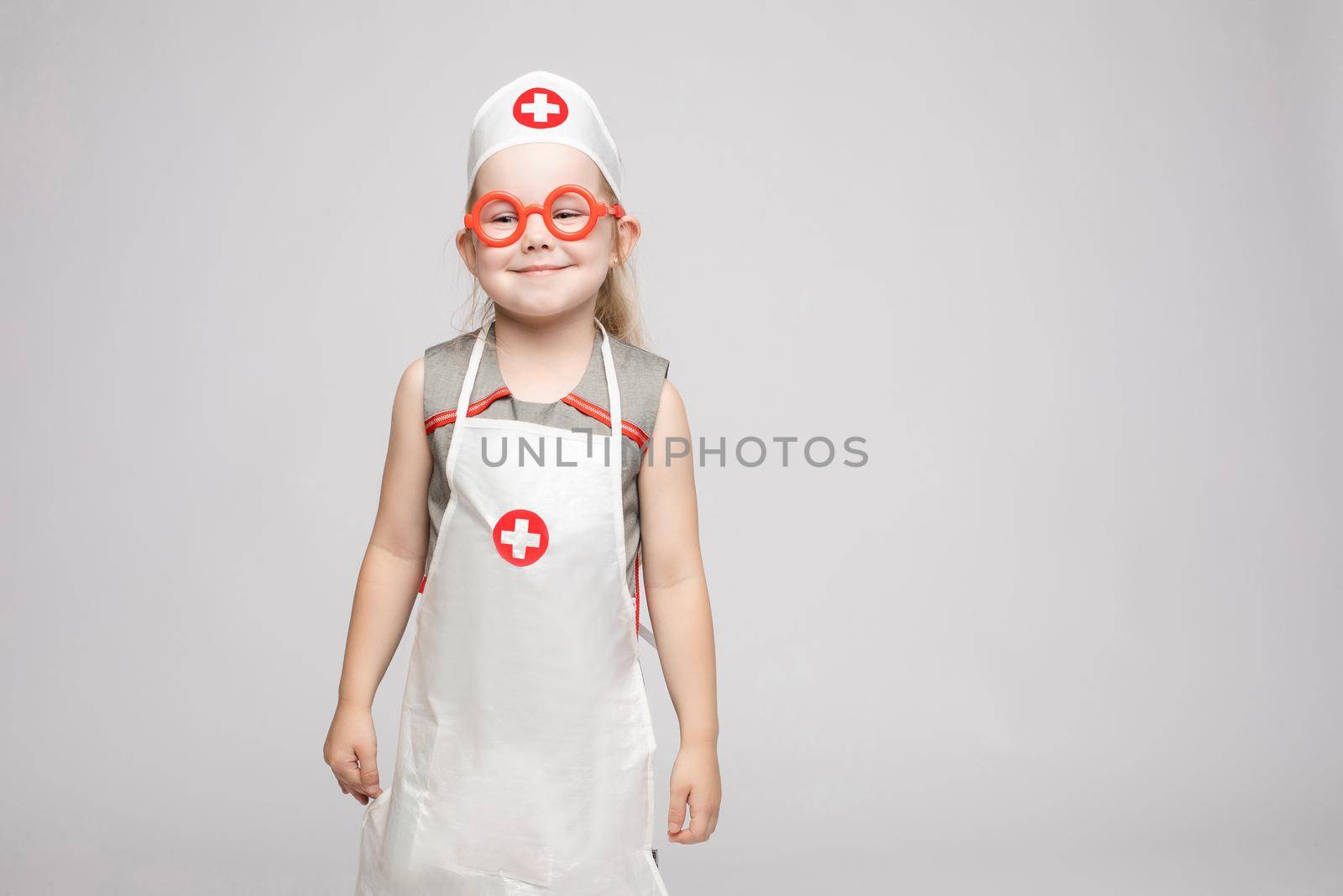 Stock photo of lovely little girl in white apron and a hat playing in a doctor. She is pointing at her hat with white cross in red circle. She is a doctor.