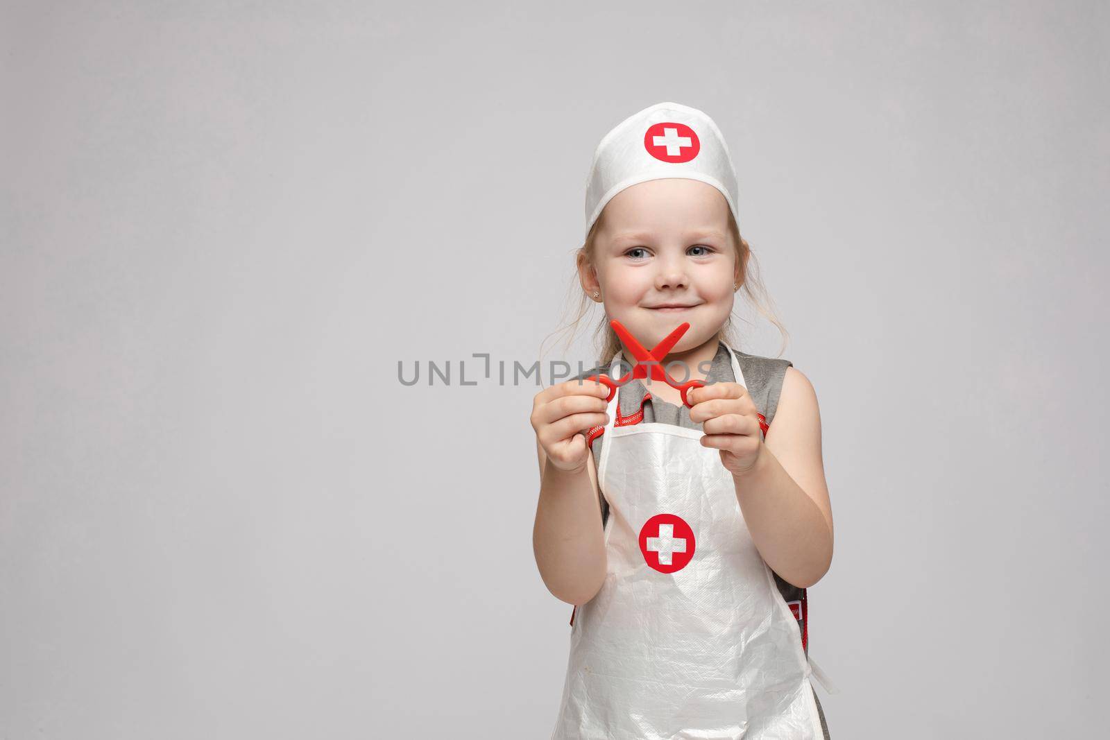 Stock photo portrait of cute little girl in doctor s hat and apron playing with plastic scissors. She is playing in doctor wearing medical uniform. Smiling at camera.