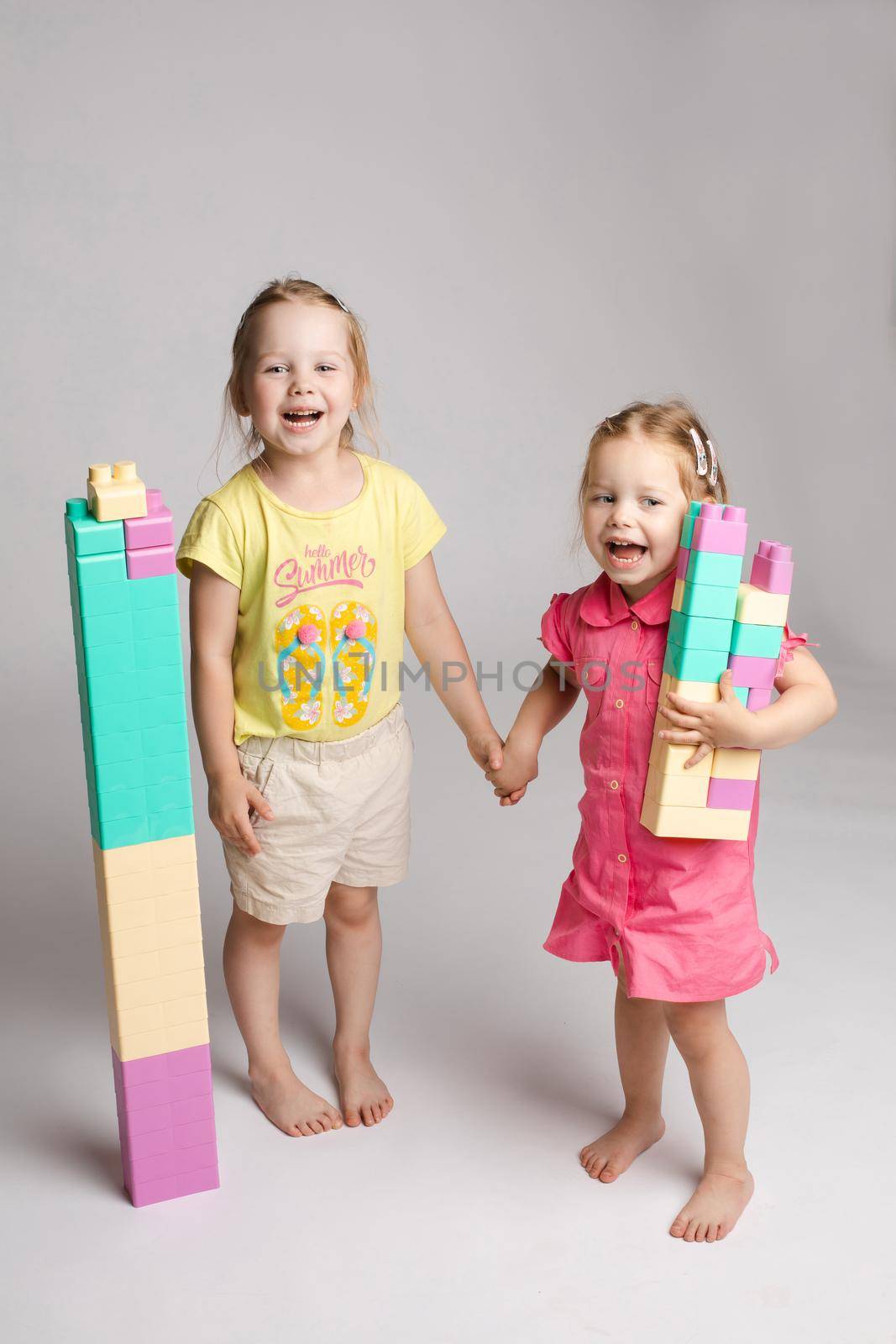 Front view of pretty sisters keeping hands together, looking at camera and smiling on isolated background. Lovely girls playing with building toys and enjoying game. Concept of happiness and joy.