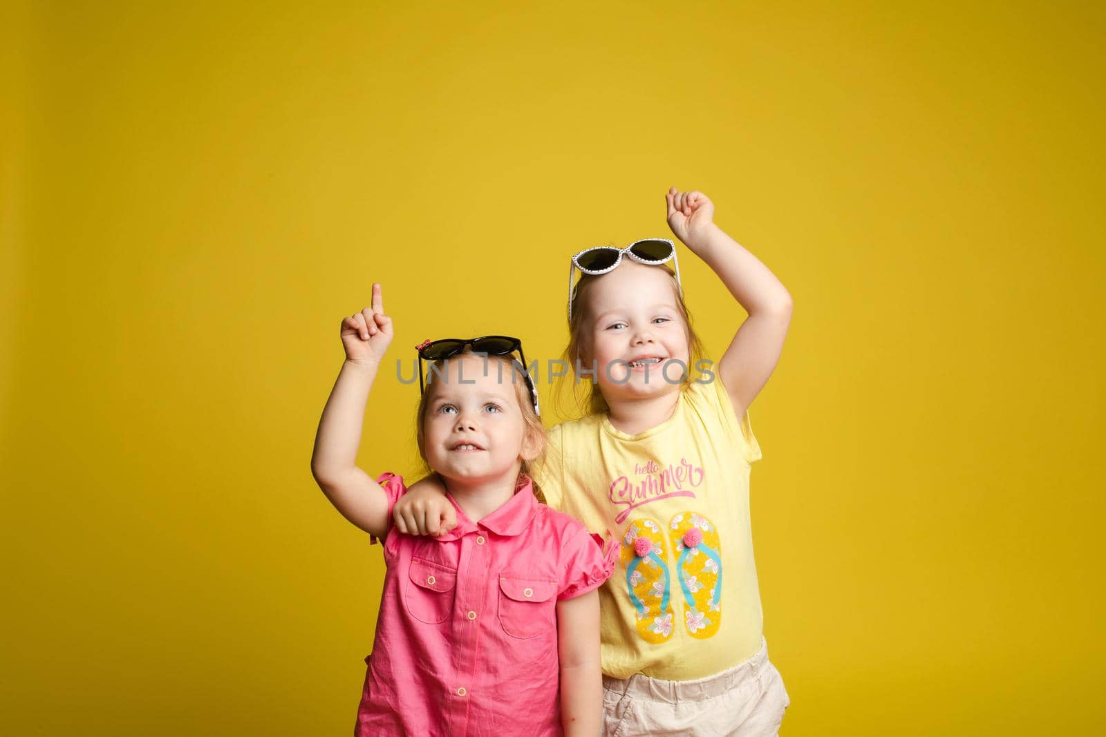 Two happy beautiful stylish little girl wearing sunglasses posing isolated yellow studio background by StudioLucky
