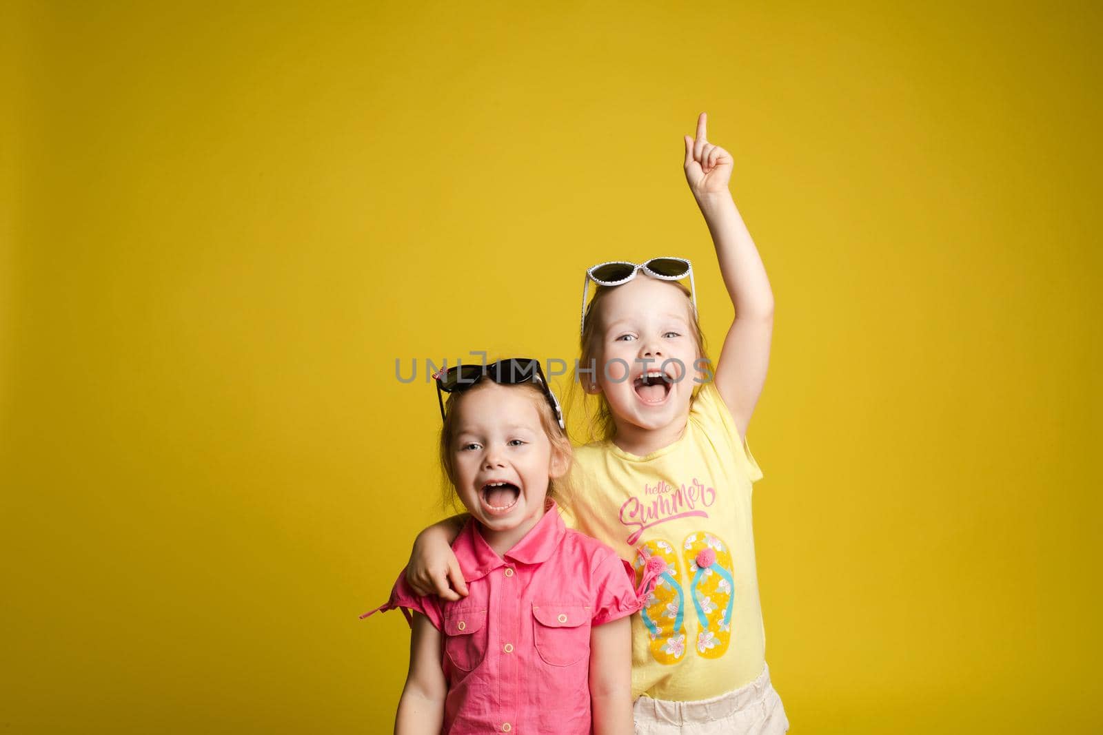 Two happy beautiful stylish little girl wearing sunglasses posing isolated at yellow studio background medium shot. Adorable laughing female child friend enjoying friendship having positive emotion