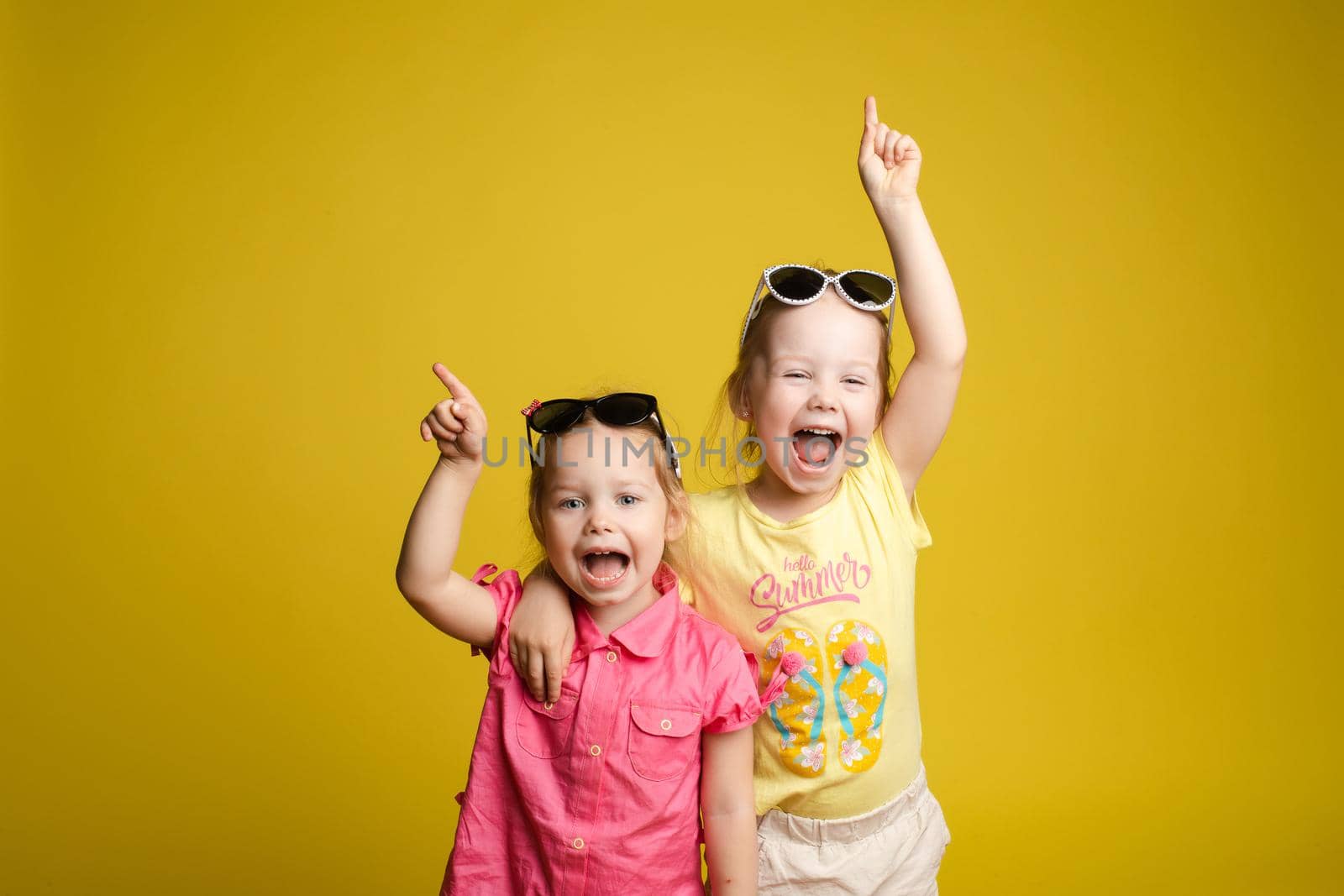 Two happy beautiful stylish little girl wearing sunglasses posing isolated at yellow studio background medium shot. Adorable laughing female child friend enjoying friendship having positive emotion