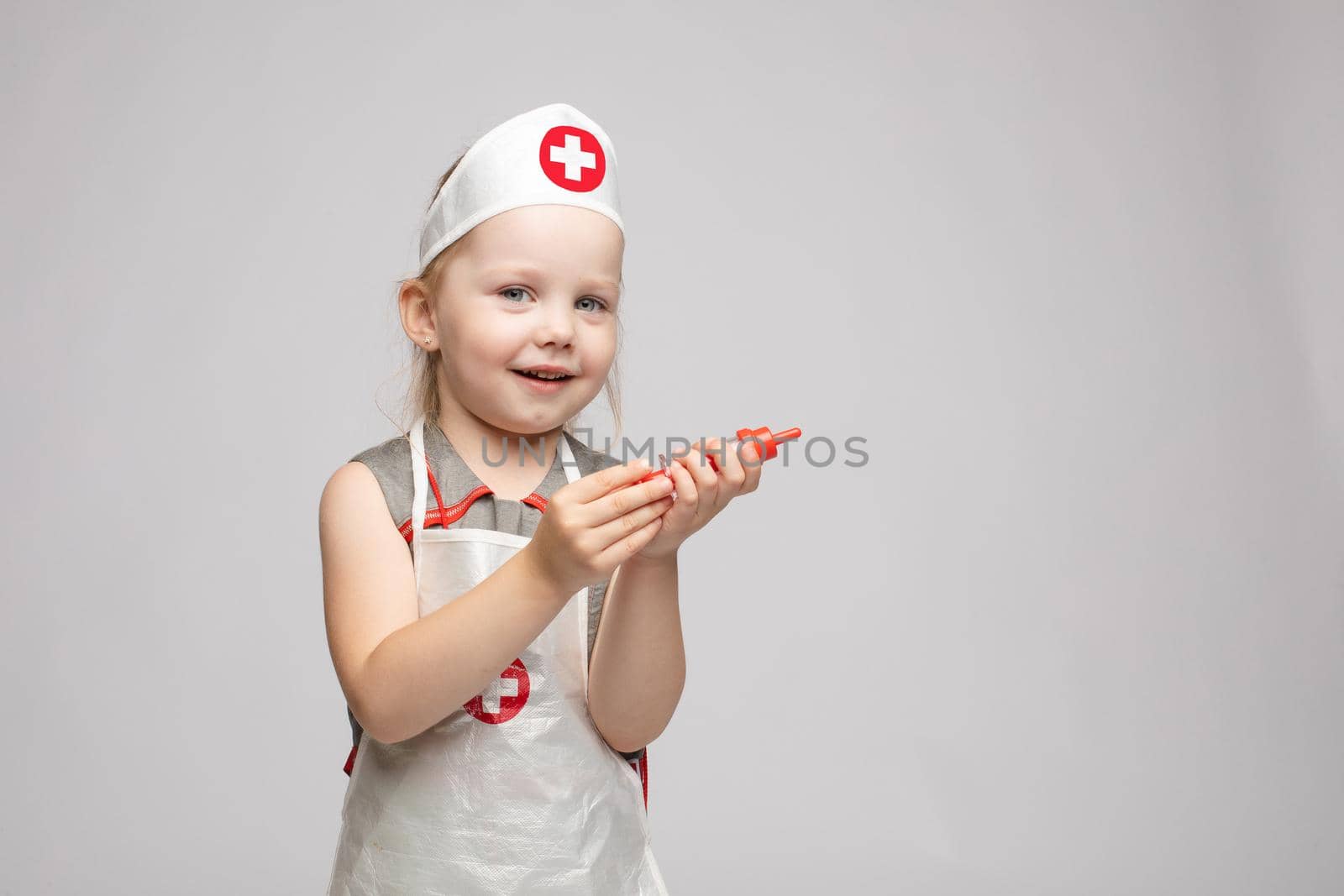 Little cute funny girl playing wearing doctor uniform holding toy syringe looking at camera. Smiling baby female child in glasses posing in nurse garb at white studio background medium long shot