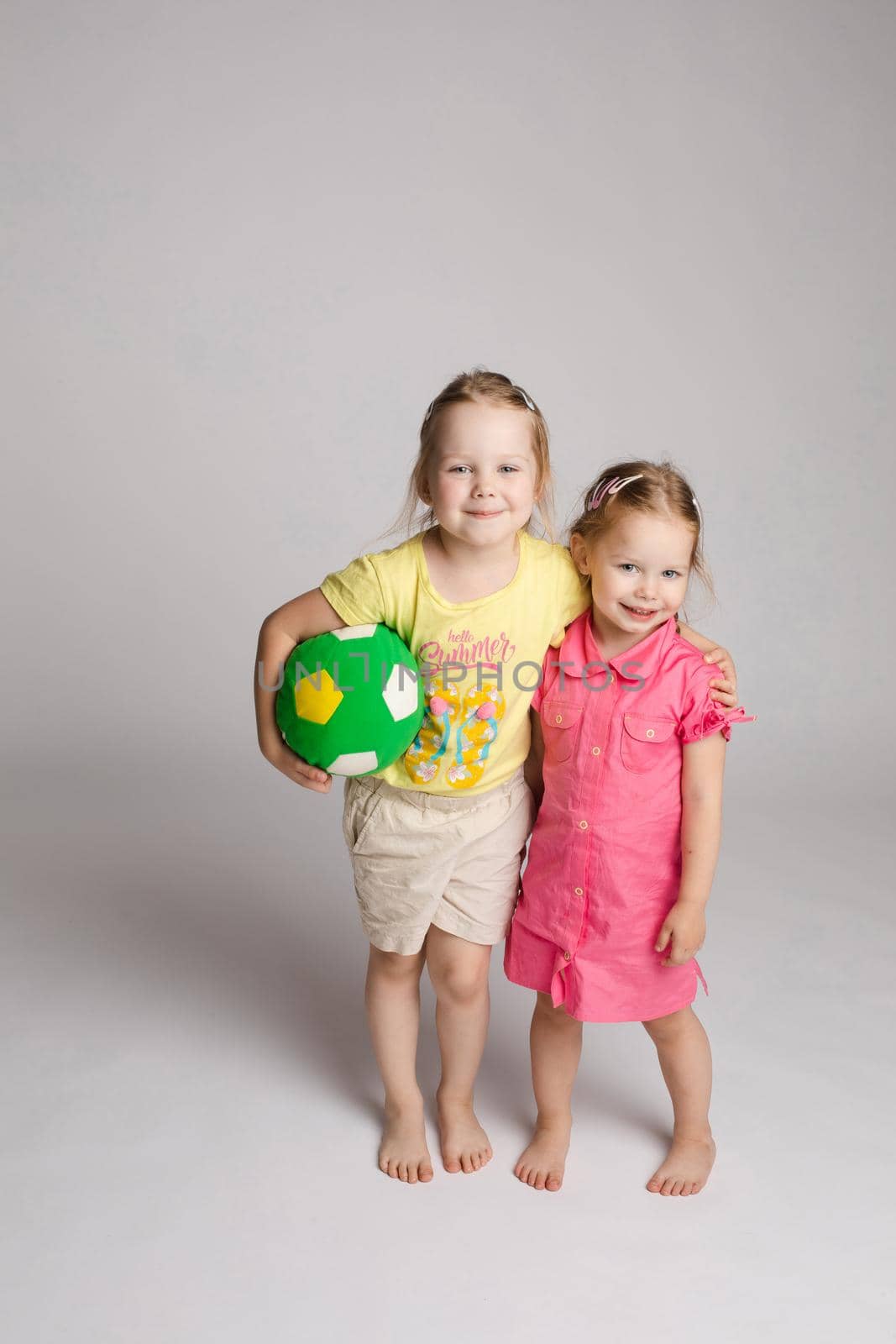 Front view of two little sisters looking at camera and posing while playing with ball. Lovely girl embracing each other, keeping toy and smiling on isolated background. Concept of joy and childhood.