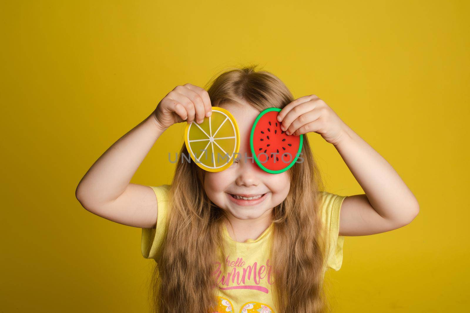 Front view of happy girl standing on yellow isolated background and closing eyes with toys. Cheerful longhaired child laughing and playing hide and seek. Concept of game and happiness.