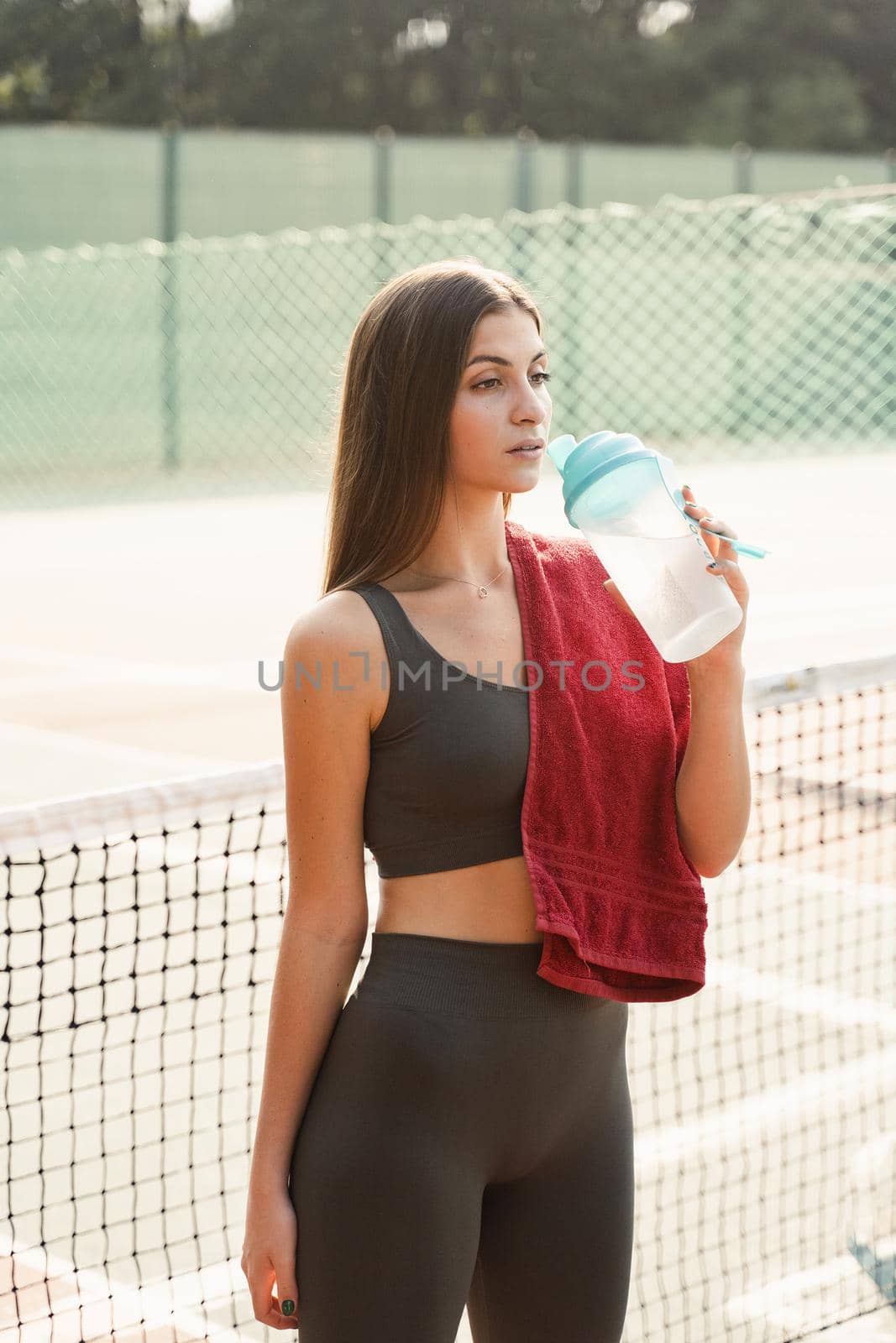 Fit sports model is resting after training and drinking water from a bottle. Sports lifestyle. Beautiful girl posing on the tennis court