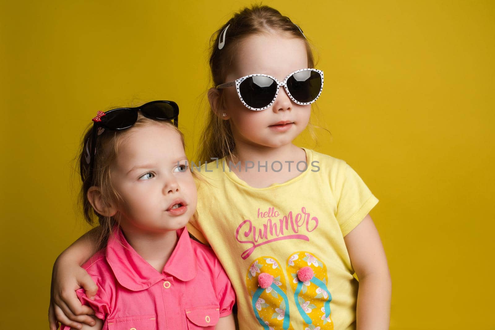 Two happy beautiful stylish little girl wearing sunglasses posing isolated at yellow studio background medium shot. Adorable laughing female child friend enjoying friendship having positive emotion