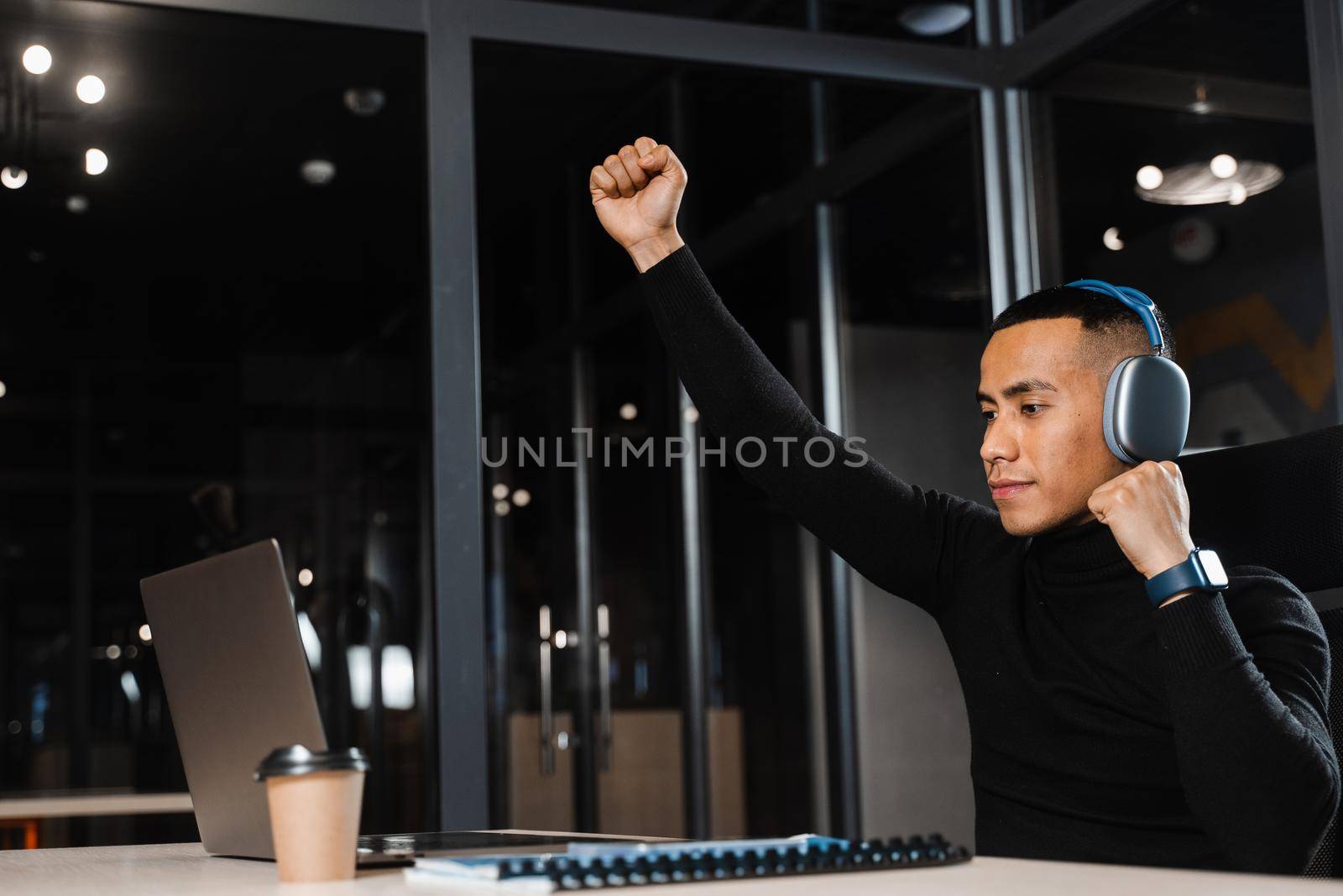Asian man raises his hands and win bet online and earn money. Happy filipino guy with laptop and headphones celebrating victory in game.