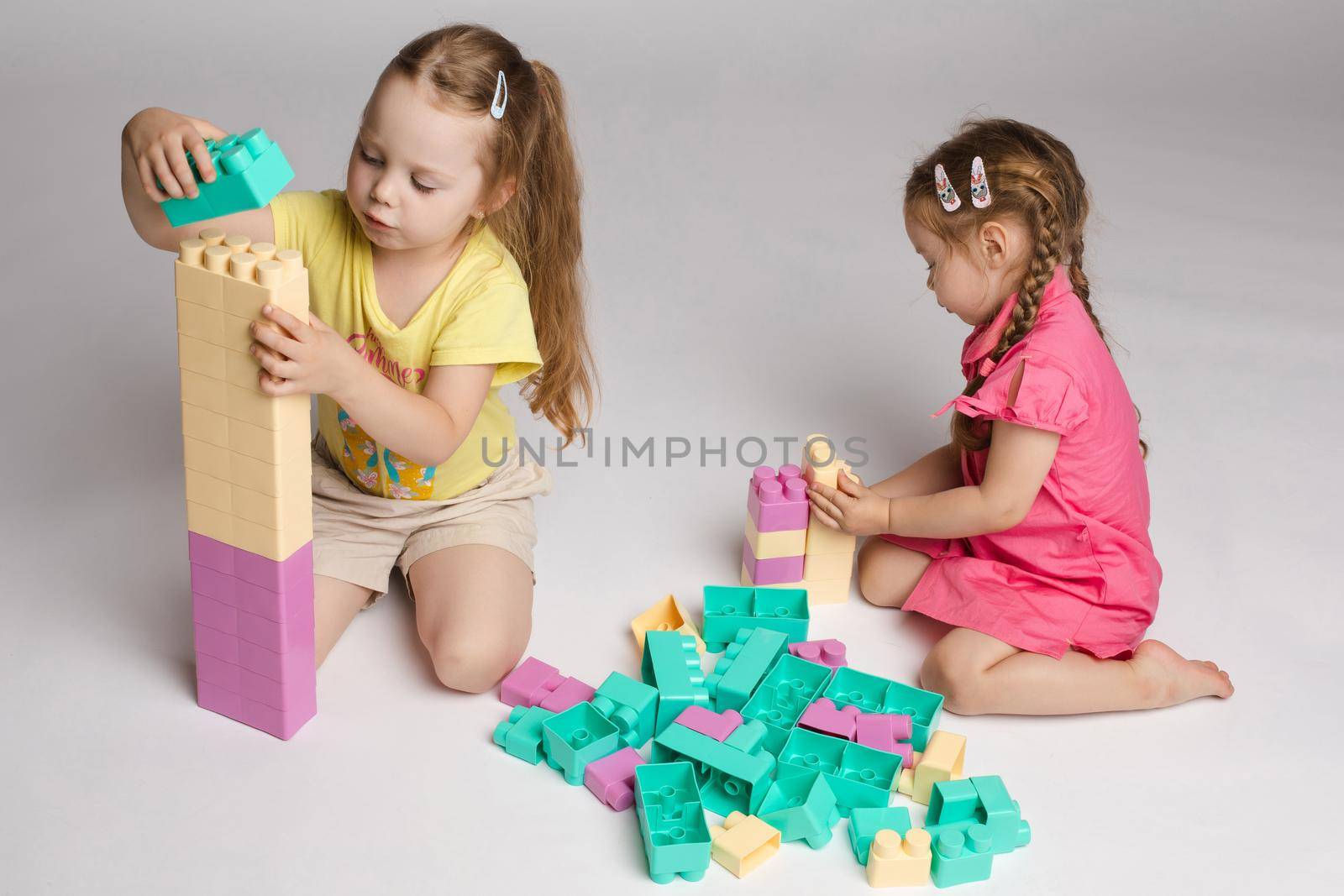 View from side of two cute girls sitting on floor and playing with building blocks in studio. Little children enjoying game, talking and smiling on isolated background. Concept of joy and activity.