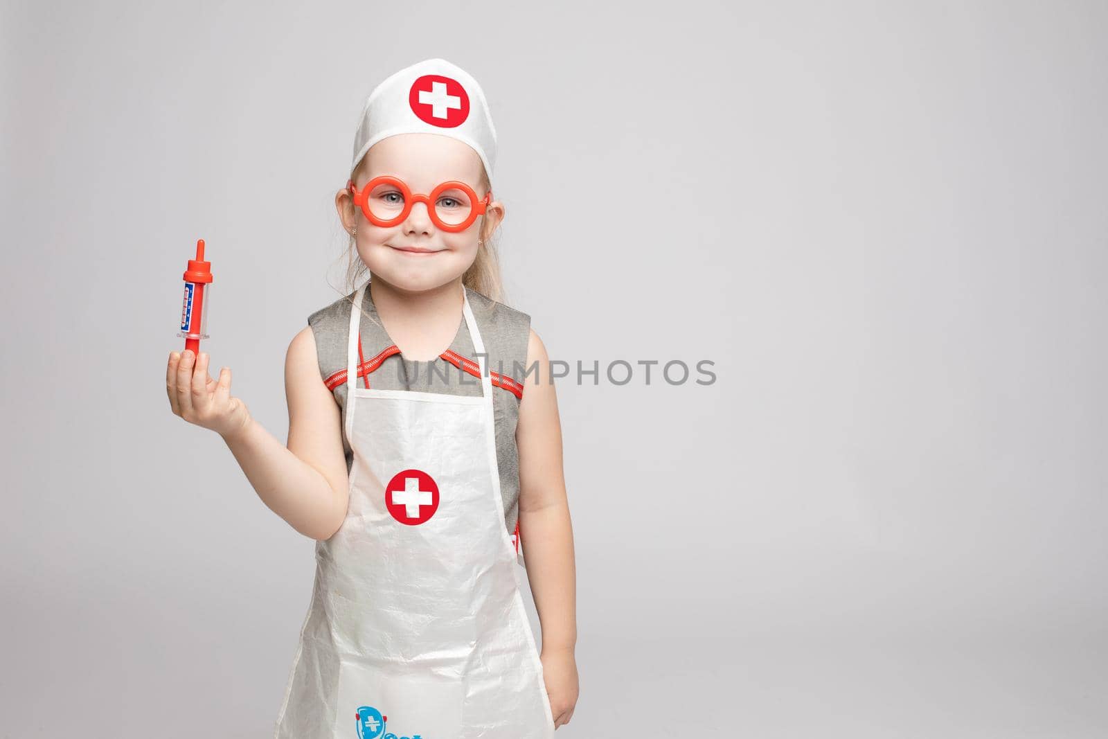 Little cute funny girl playing wearing doctor uniform holding toy syringe looking at camera. Smiling baby female child in glasses posing in nurse garb at white studio background medium long shot