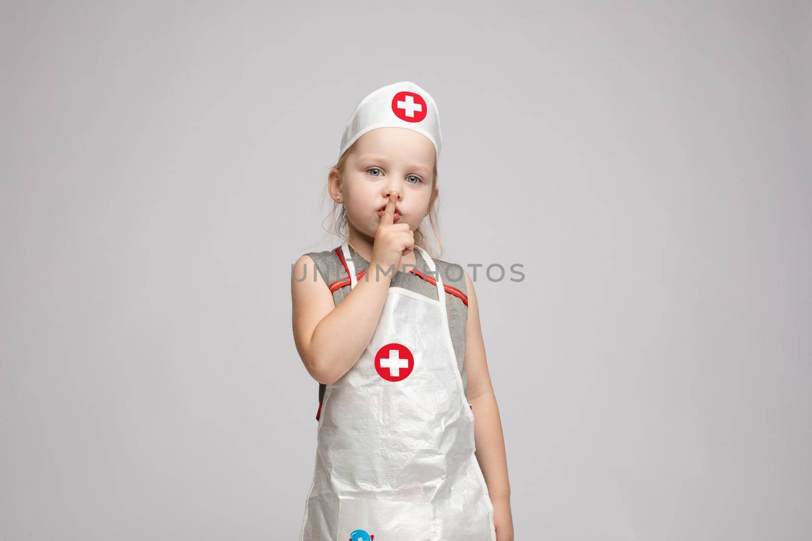 Cropped view of funny little girl wearing doctor uniform looking at camera and shoring sign of silence. Preschool child playing nurse and enjoying game on isoalted background. Concept of fun and joy.