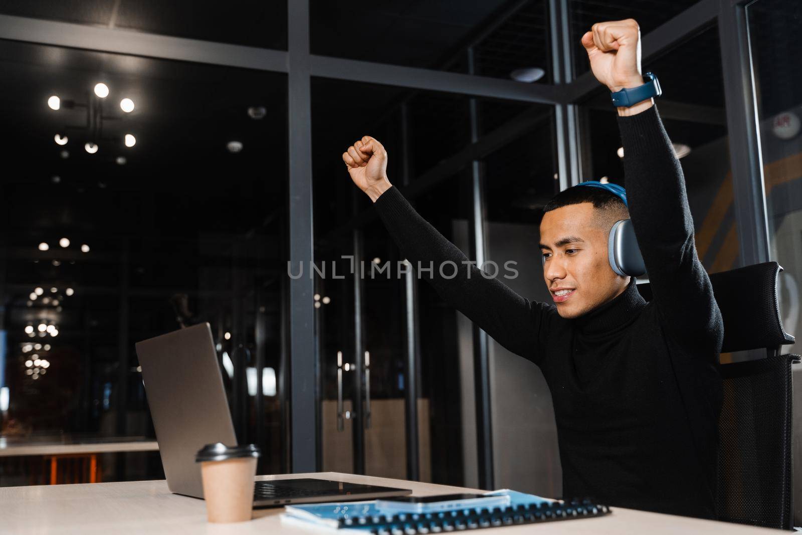 Asian man raises his hands and win bet online and earn money. Happy filipino guy with laptop and headphones celebrating victory in game by Rabizo