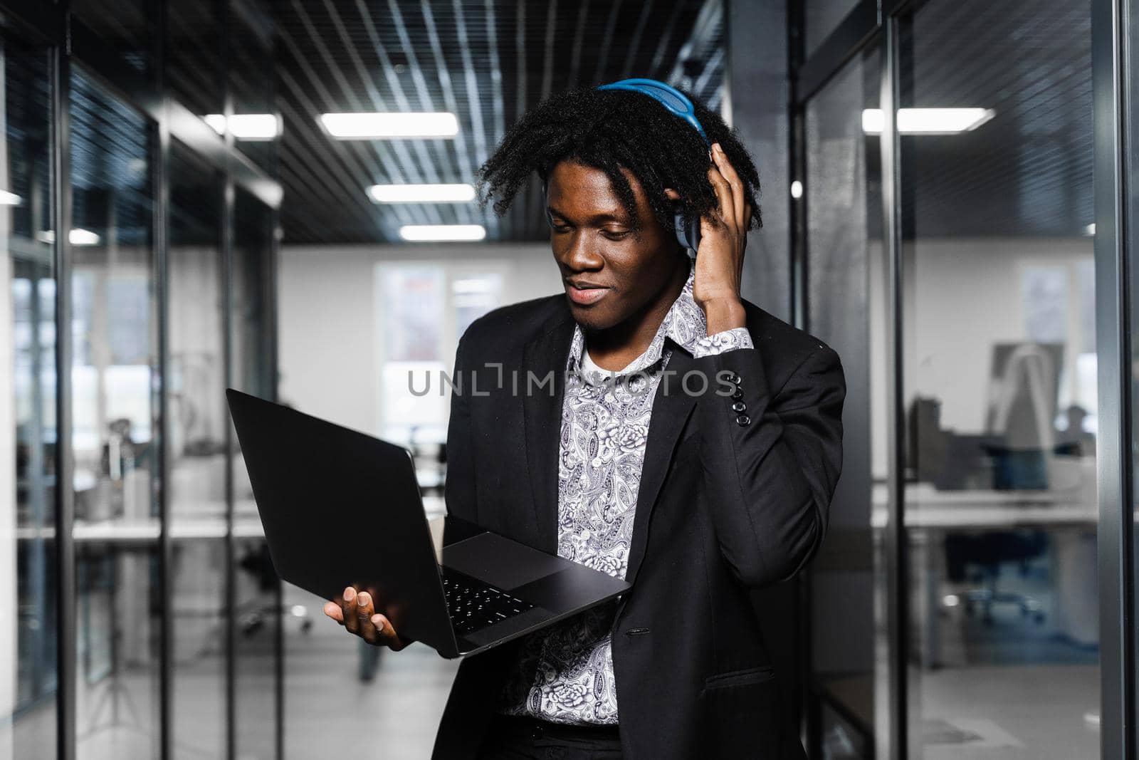 Cheerful black man in headphones listen audio podcast and enjoy music. Handsome african man with laptop in the office. Party concept. by Rabizo