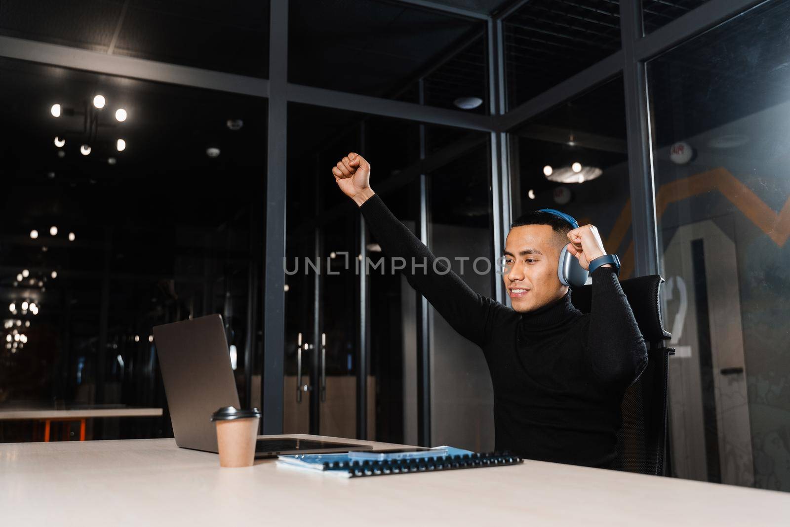 Asian man raises his hands and win bet online and earn money. Happy filipino guy with laptop and headphones celebrating victory in game.