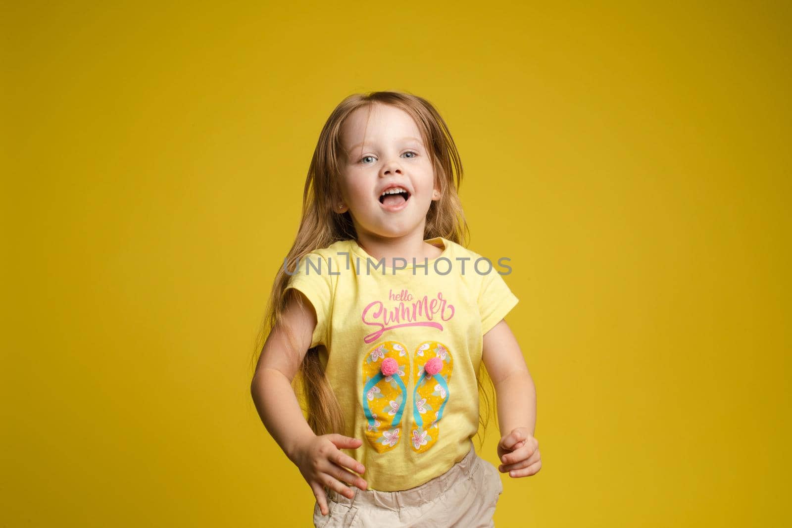 Front view of beautiful longhaired girl in cute shirt playing with hair and twirling in studio. Sunny little child looking at camera and posing on yellow isolated background. Concept of childhood.