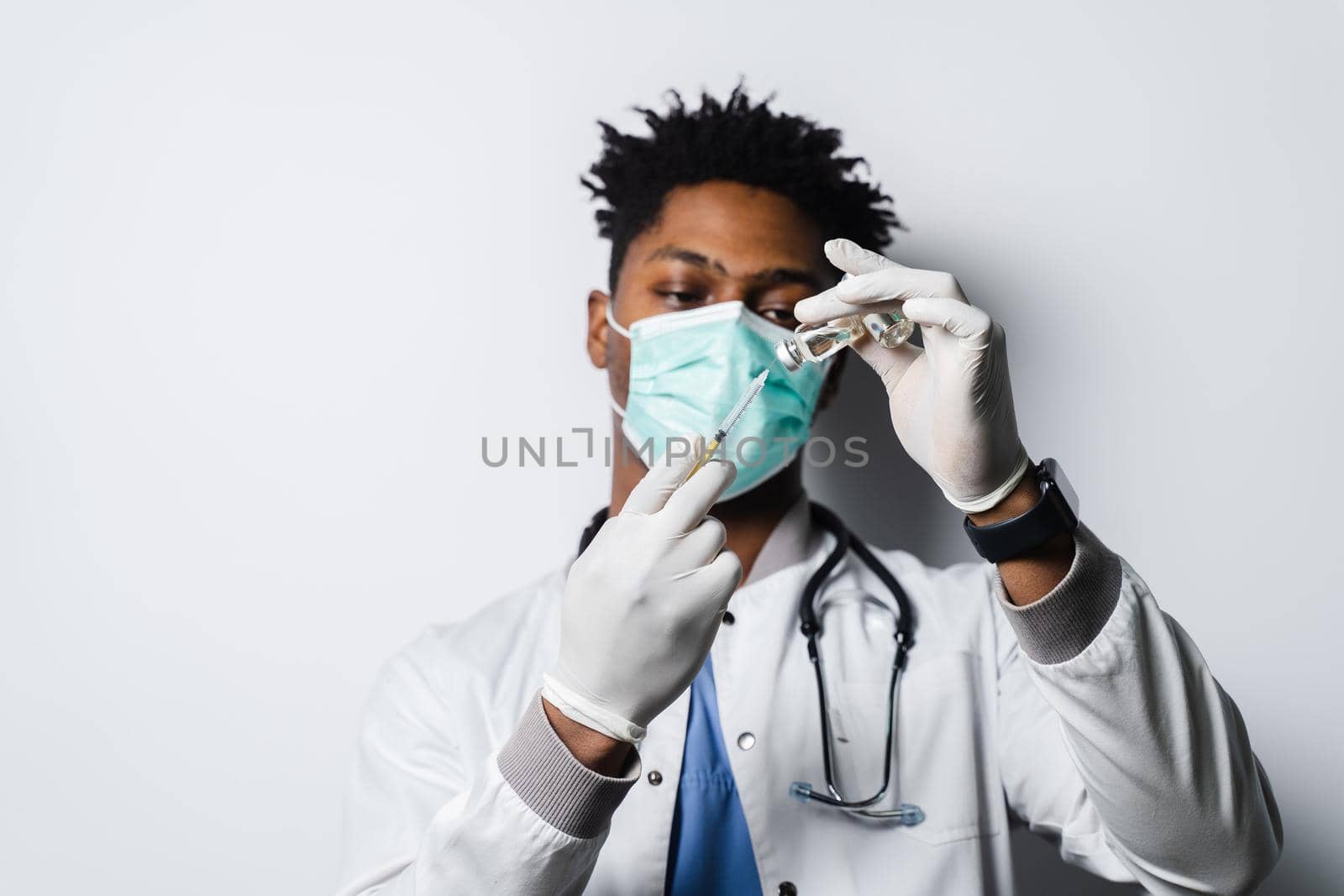 African doctor in a medical mask prepares to give an injection of a coronavirus covid-19 vaccine. Black man in medical mask is making vaccination on white background. by Rabizo