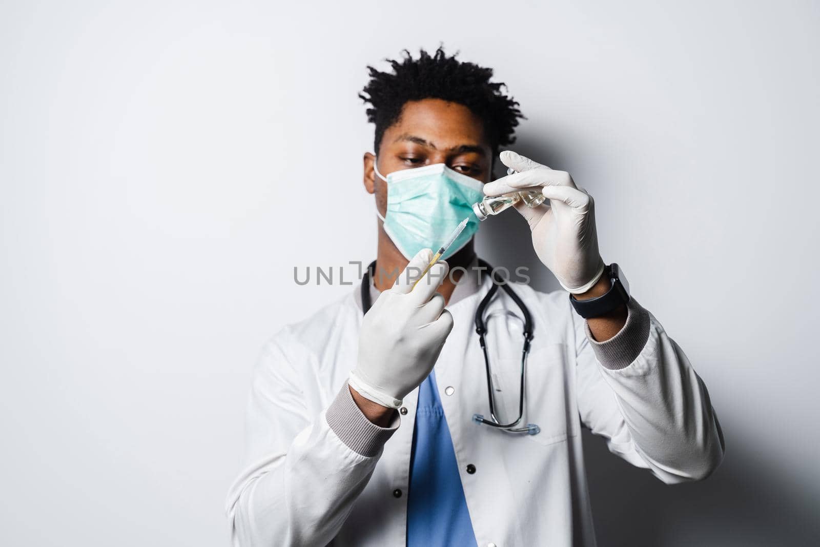 African doctor in a medical mask prepares to give an injection of a coronavirus covid-19 vaccine. Black man in medical mask is making vaccination on white background. by Rabizo