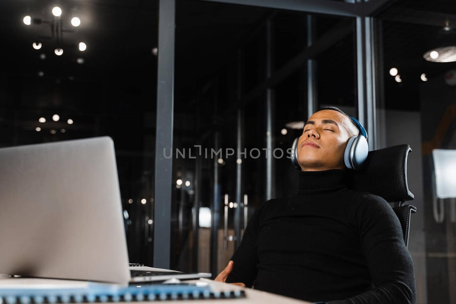 Overtime working. Asian man with closed eyes is resting at work at laptop. Tired male programmer taking a break due to overtime work.