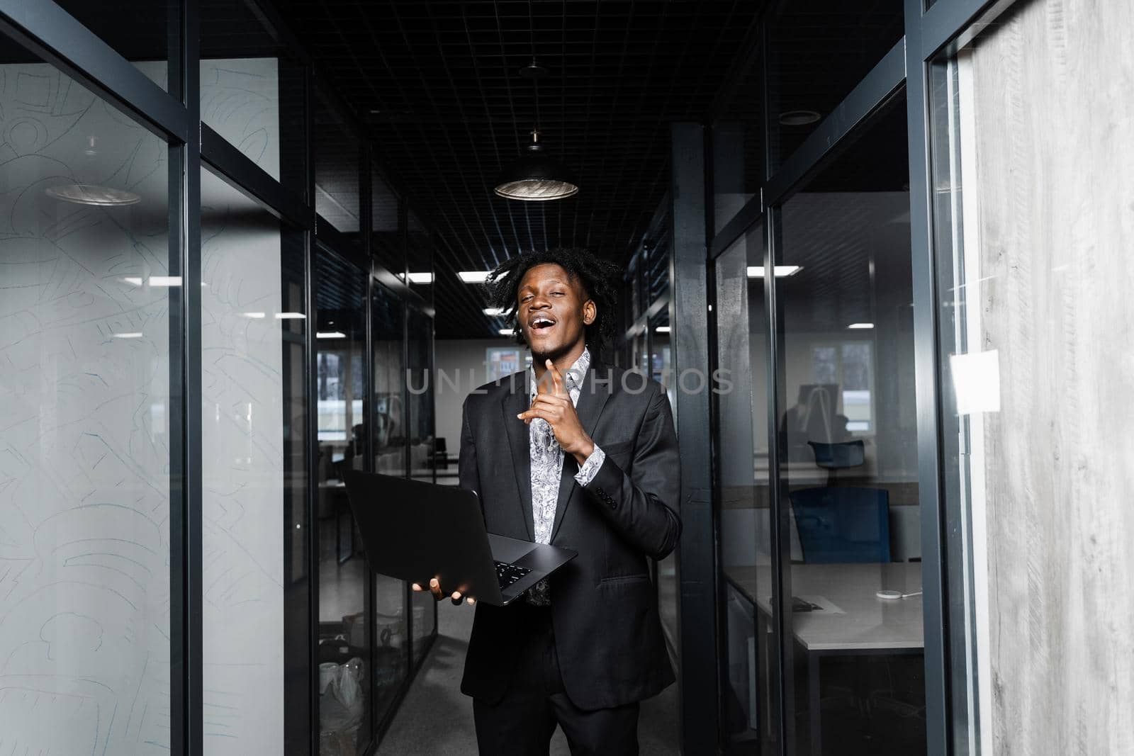 Cheerful black business man with laptop is pointing fingers up and smiling in office