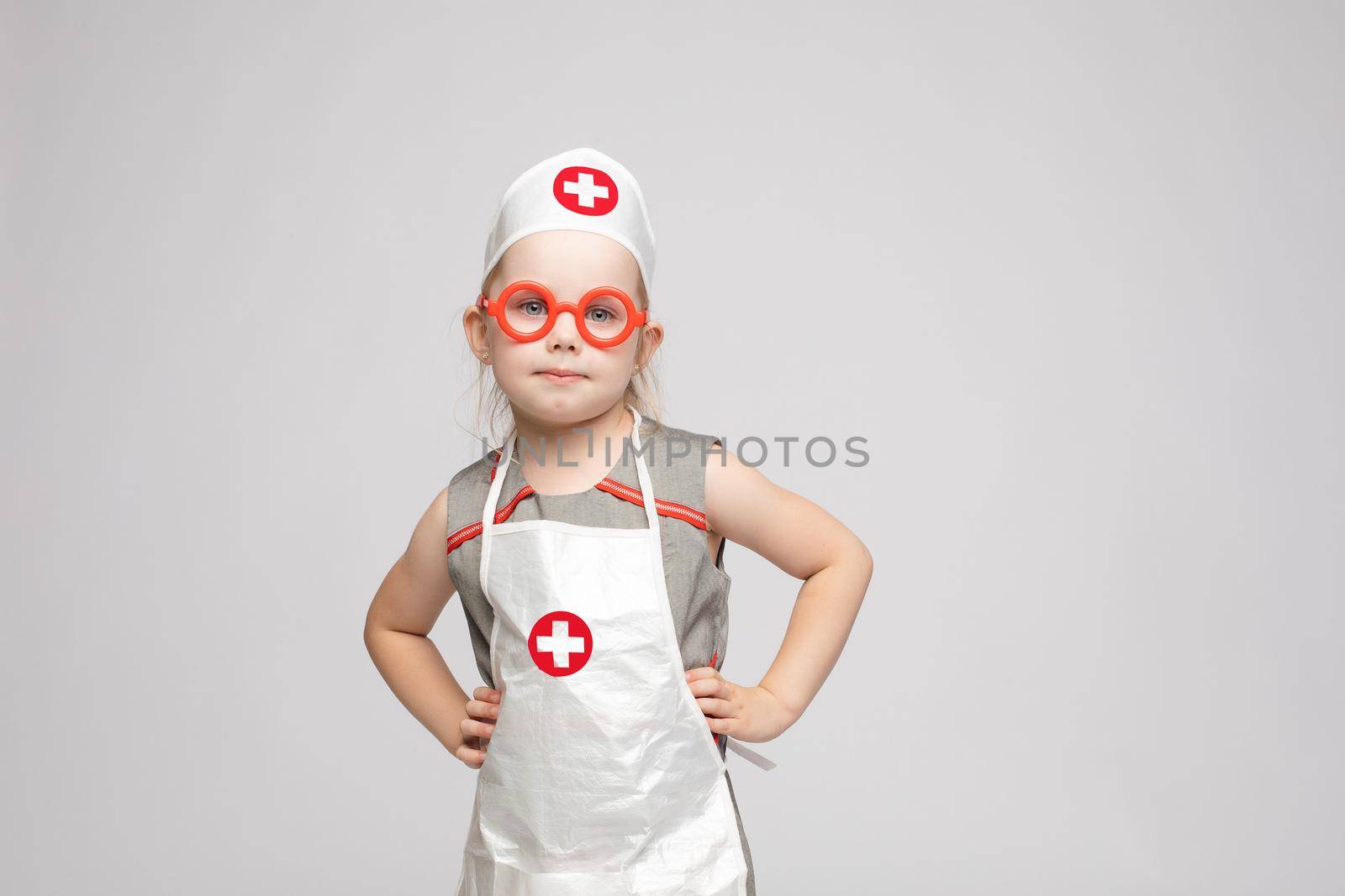 Stock photo of lovely little girl in white apron and a hat playing in a doctor. She is pointing at her hat with white cross in red circle. She is a doctor.