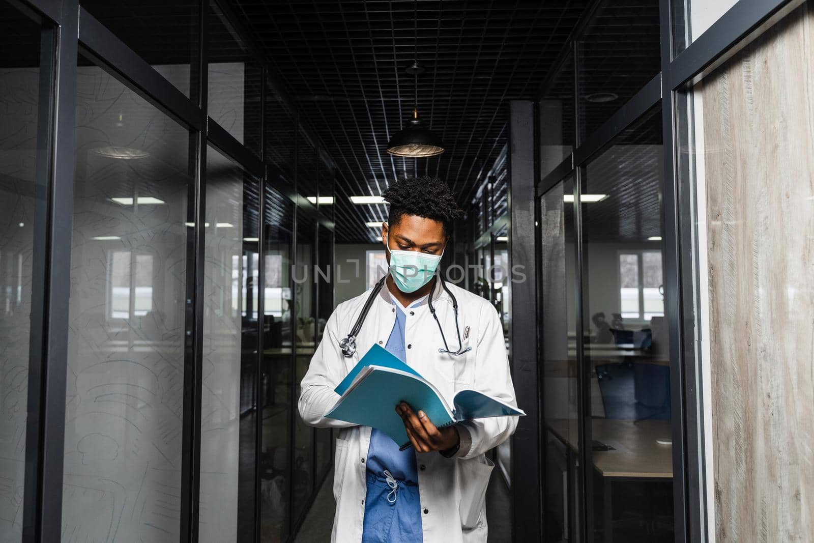 Black medical student reads books and prepares for an exam. African in a medical robe and mask looks at the notes. by Rabizo