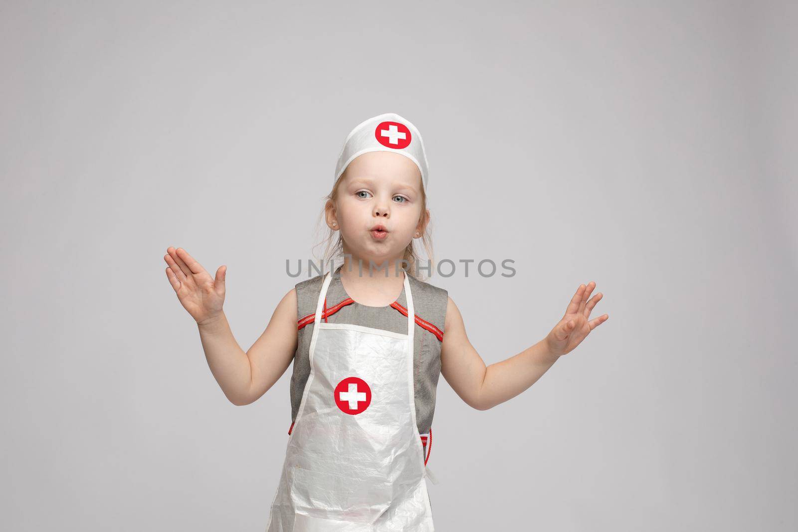 Stock photo of lovely little girl in white apron and a hat playing in a doctor. She is pointing at her hat with white cross in red circle. She is a doctor.