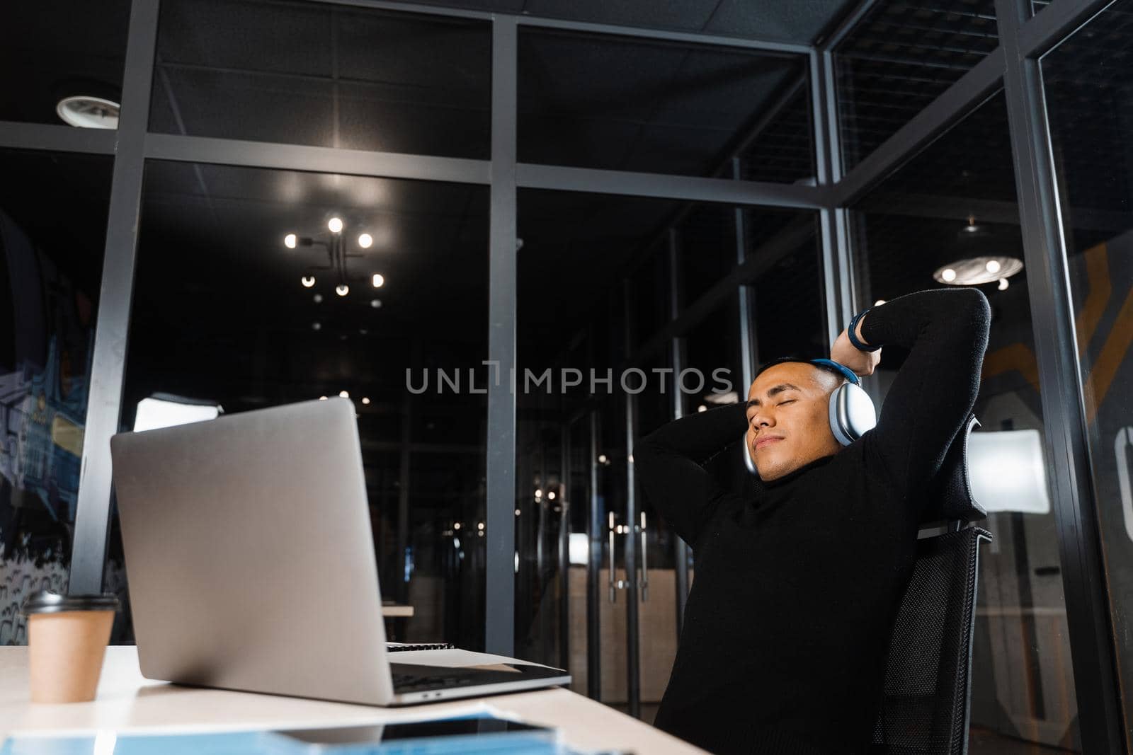 Asian man with closed eyes listening to relaxing music at work at laptop. Break at workplace for rest from overtime work. by Rabizo
