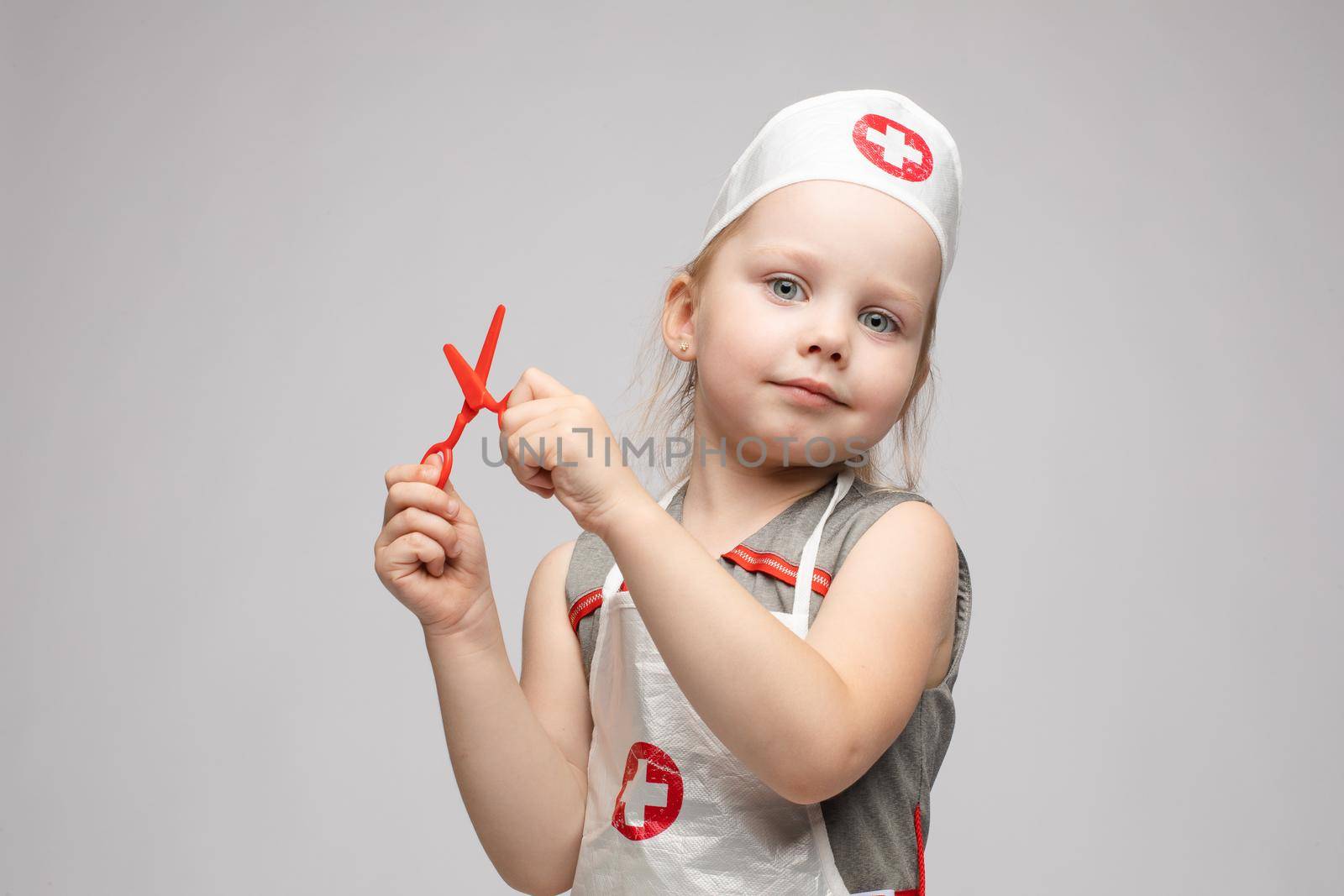 Stock photo portrait of cute little girl in doctor s hat and apron playing with plastic scissors. She is playing in doctor wearing medical uniform. Smiling at camera.