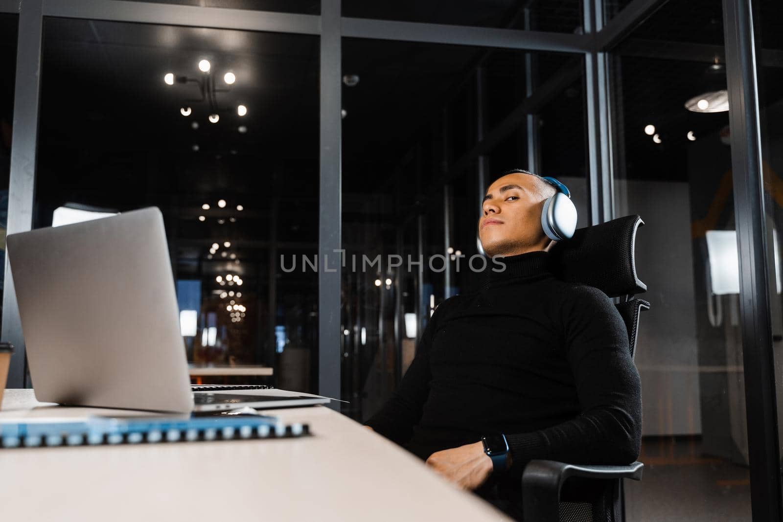 Overtime working. Asian man with closed eyes is resting at work at laptop. Tired male programmer taking a break due to overtime work by Rabizo