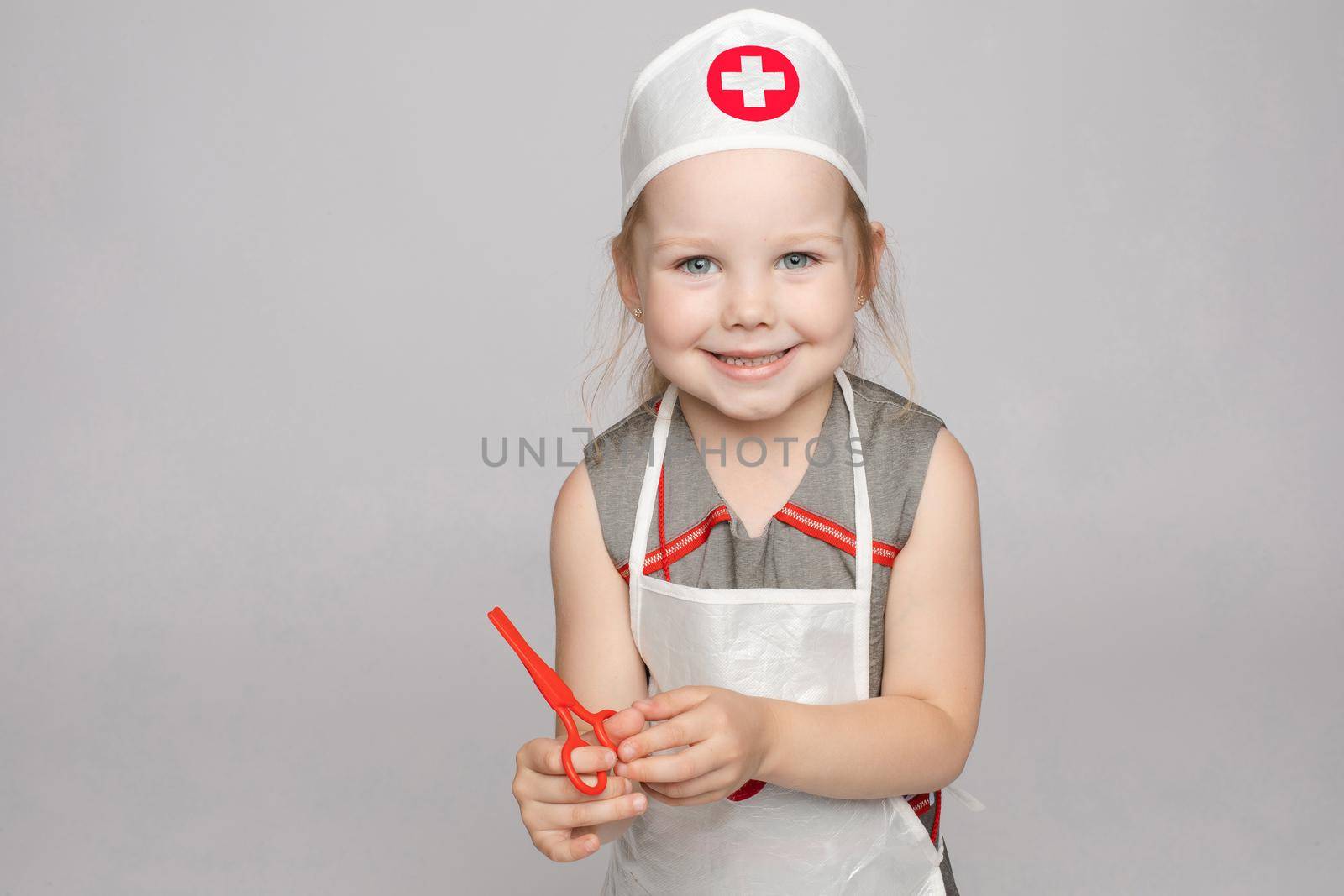 Stock photo portrait of cute little girl in doctor s hat and apron playing with plastic scissors. She is playing in doctor wearing medical uniform. Smiling at camera.