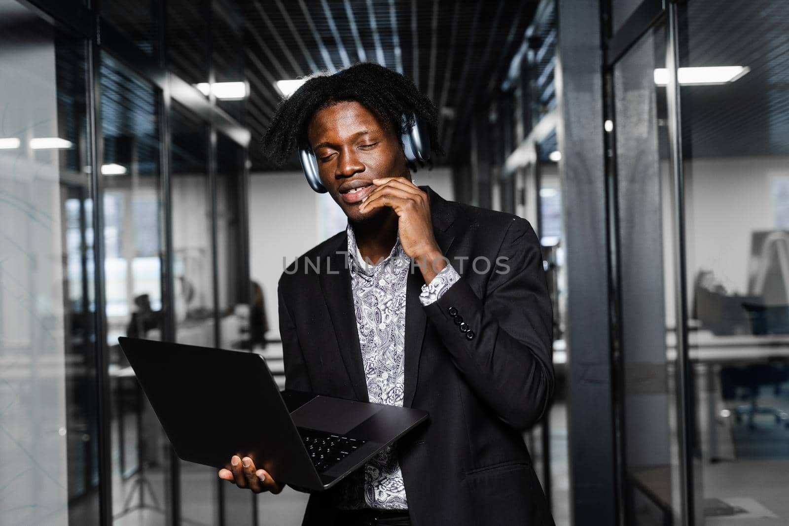 Cheerful black man in headphones listen audio podcast and enjoy music. Handsome african man with laptop in the office. Party concept. by Rabizo