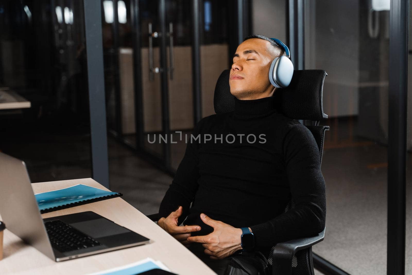 Asian man with closed eyes listening to relaxing music at work at laptop. Break at workplace for rest from overtime work. by Rabizo