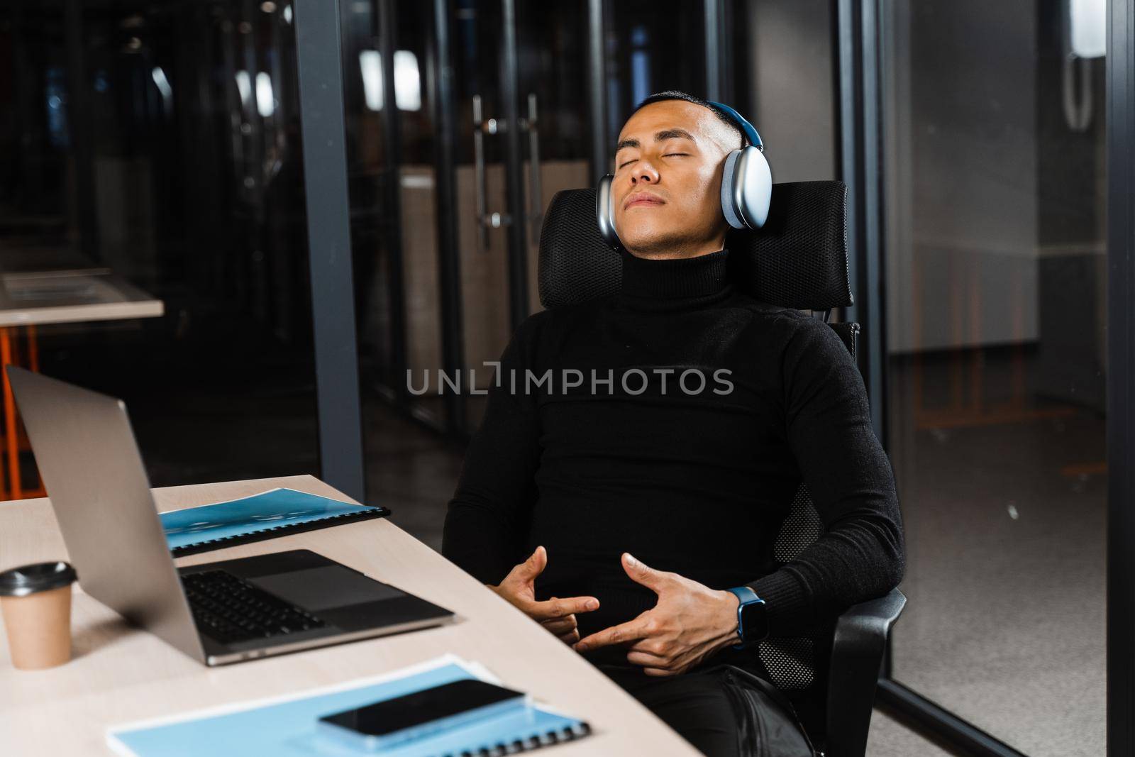 Asian man with closed eyes listening to relaxing music at work at laptop. Break at workplace for rest from overtime work