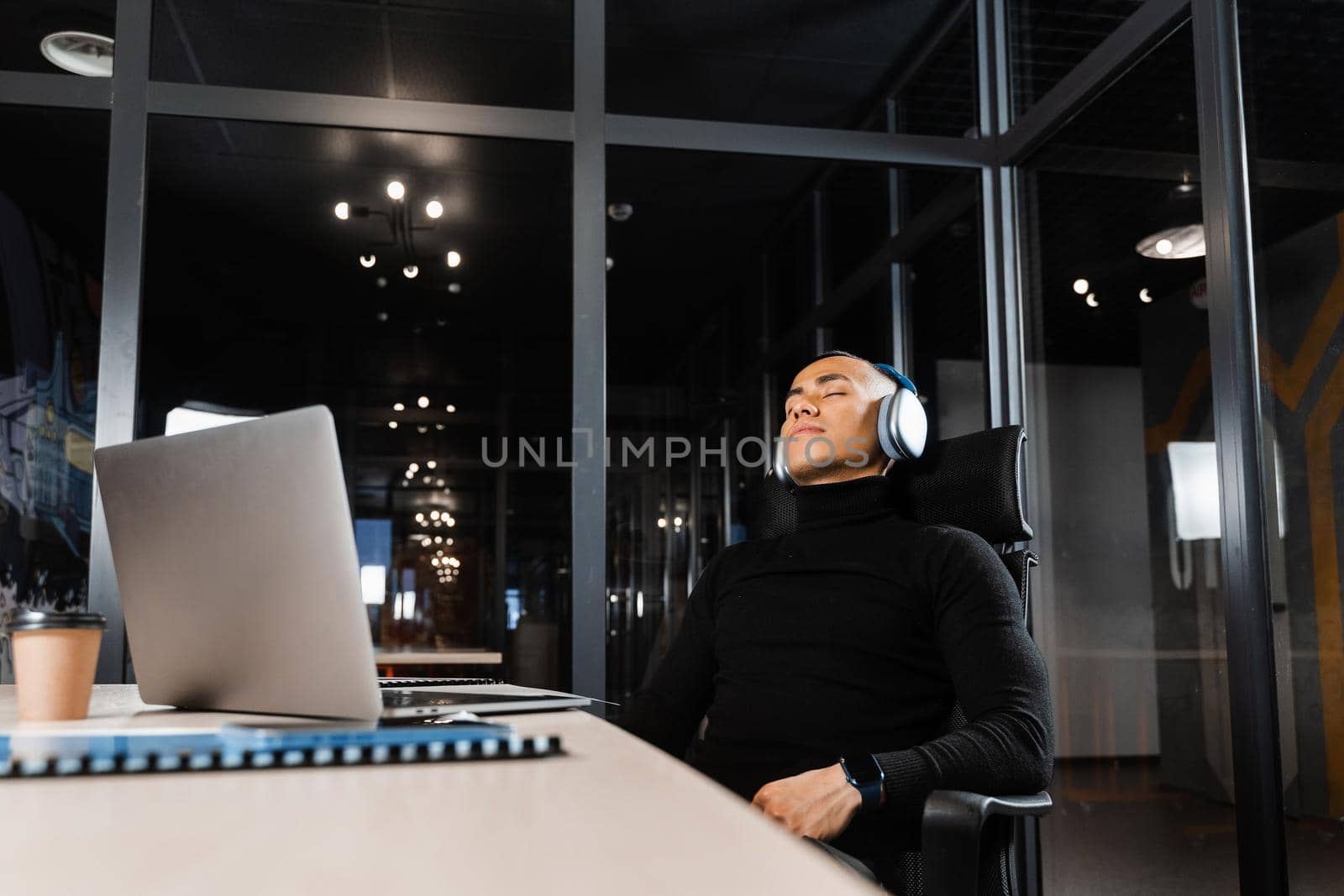 Asian man with closed eyes listening to relaxing music at work at laptop. Break at workplace for rest from overtime work